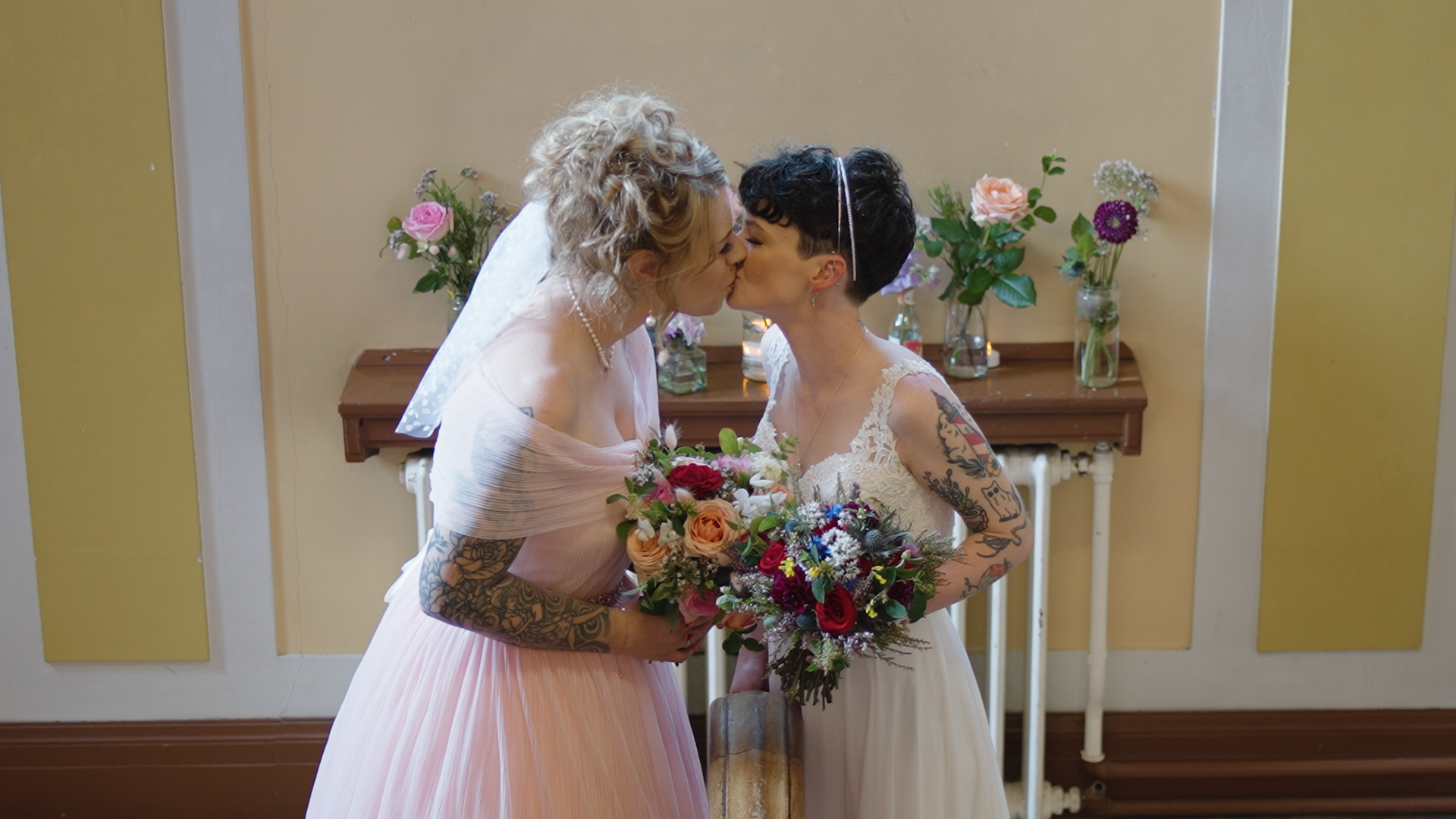 Two brides kissing, holding colorful bouquets, against a backdrop with flowers in vases.