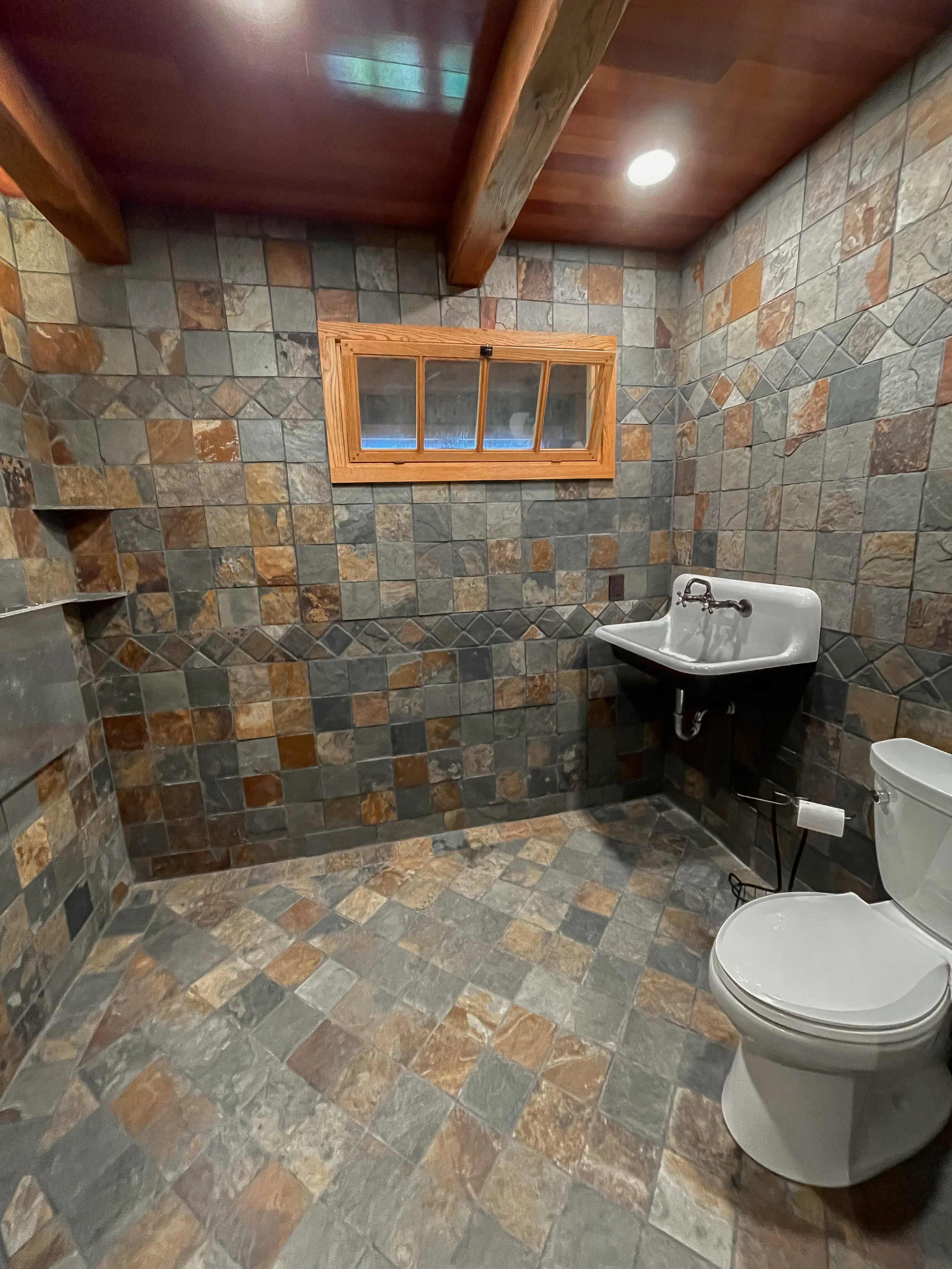 A bathroom with multicolored slate tile walls and floor, a small window with a wooden frame, a white toilet, a small white sink with exposed plumbing, and wooden ceiling beams.