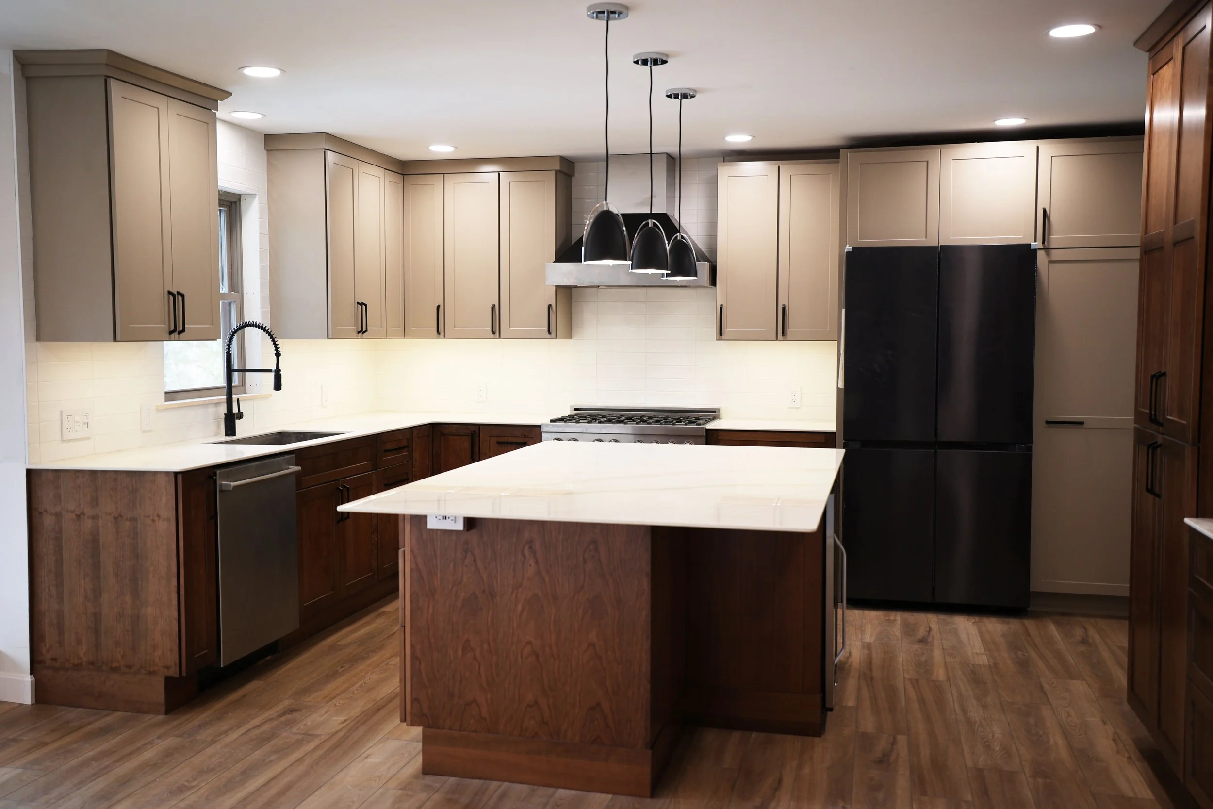 Modern kitchen with beige and dark wood cabinets, black refrigerator, white countertops, and a kitchen island, illuminated by ceiling lights and pendant lights.