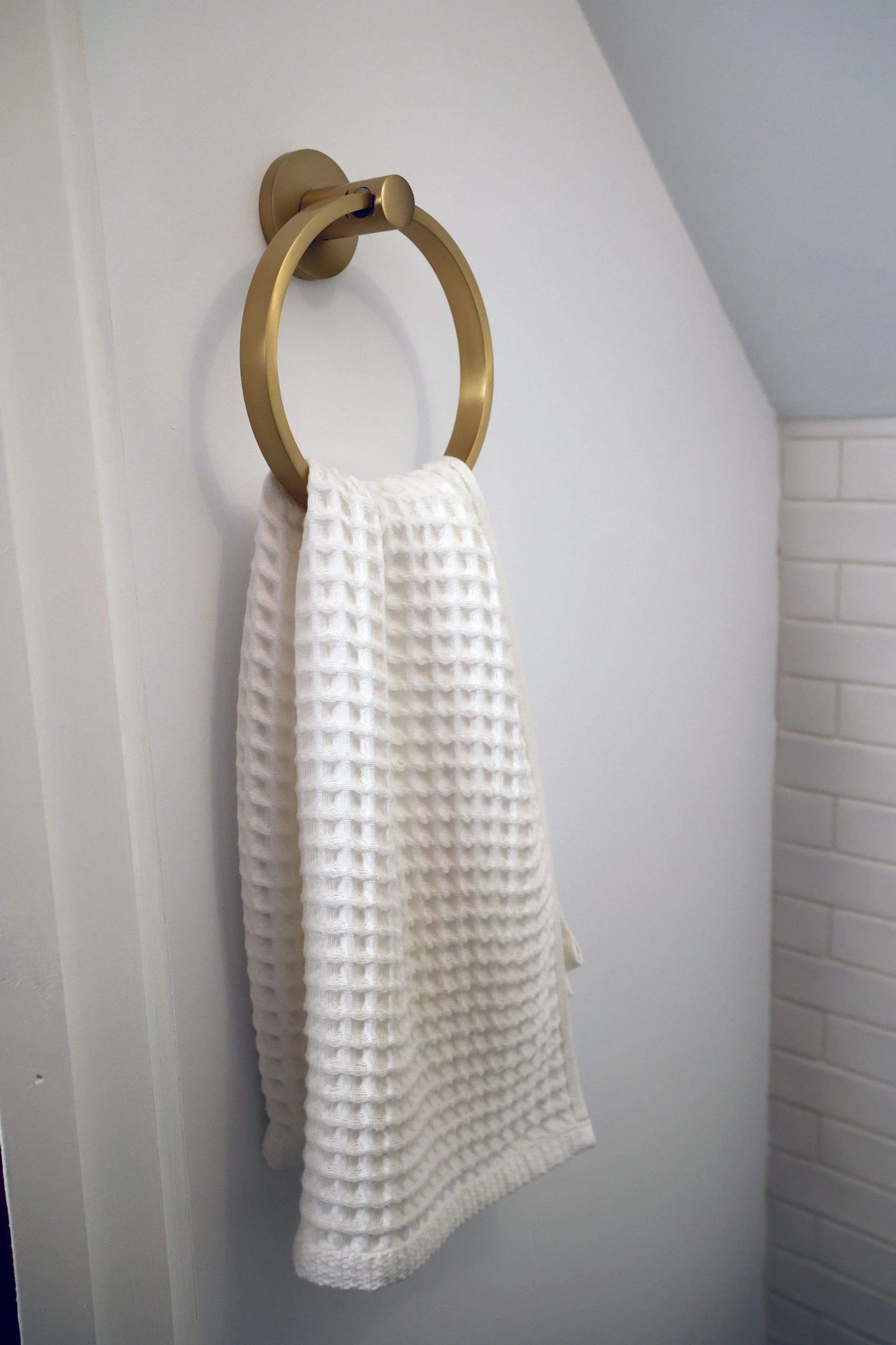 A white textured towel hanging on a gold towel ring mounted on a white wall in a bathroom.