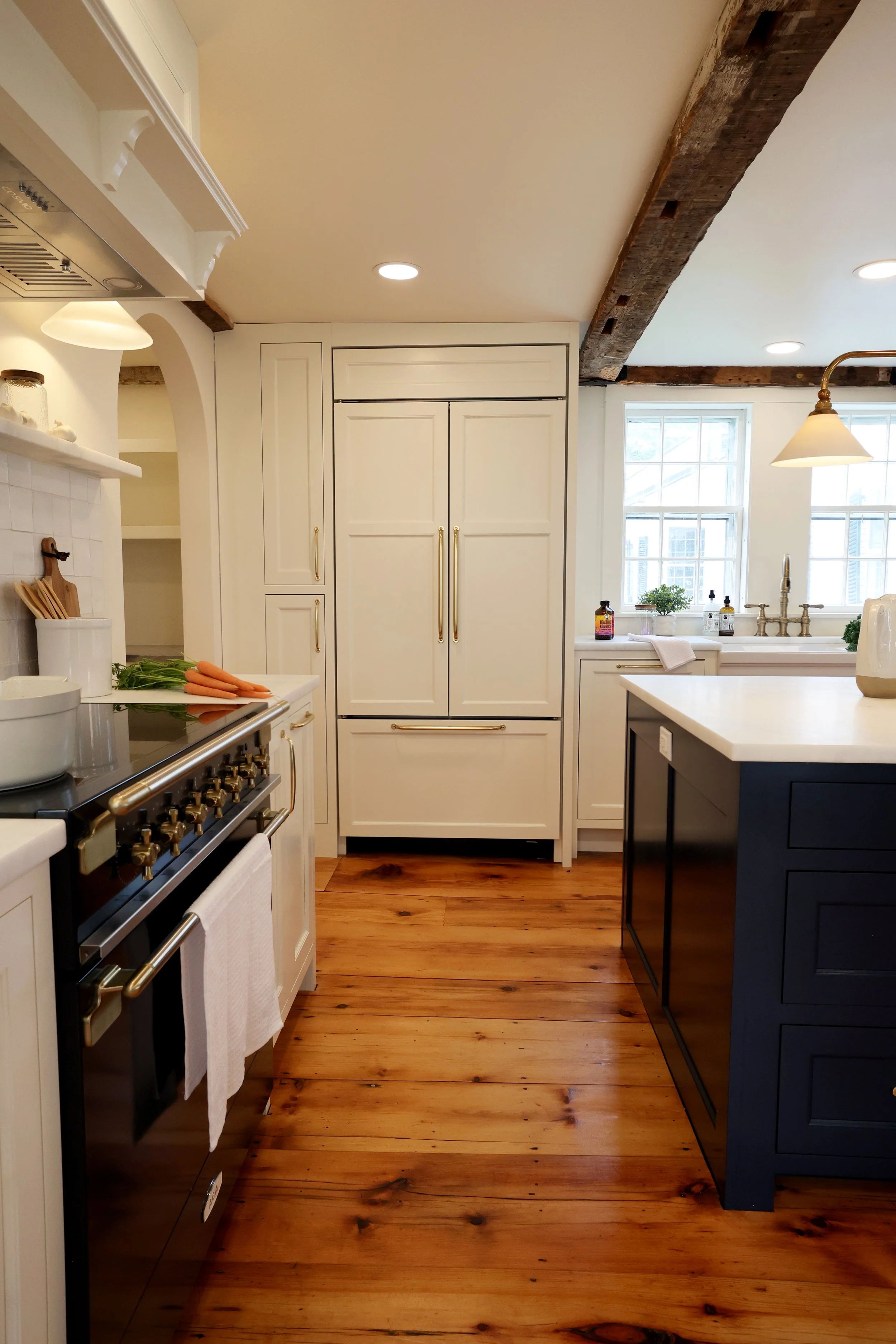 A bright kitchen with white cabinets, wooden beams on the ceiling, and a hardwood floor. There are two windows above the sink, a black island with a white countertop, and a stove with a white towel hanging from the handle. Items like carrots and green onions are on the counter near the stove.