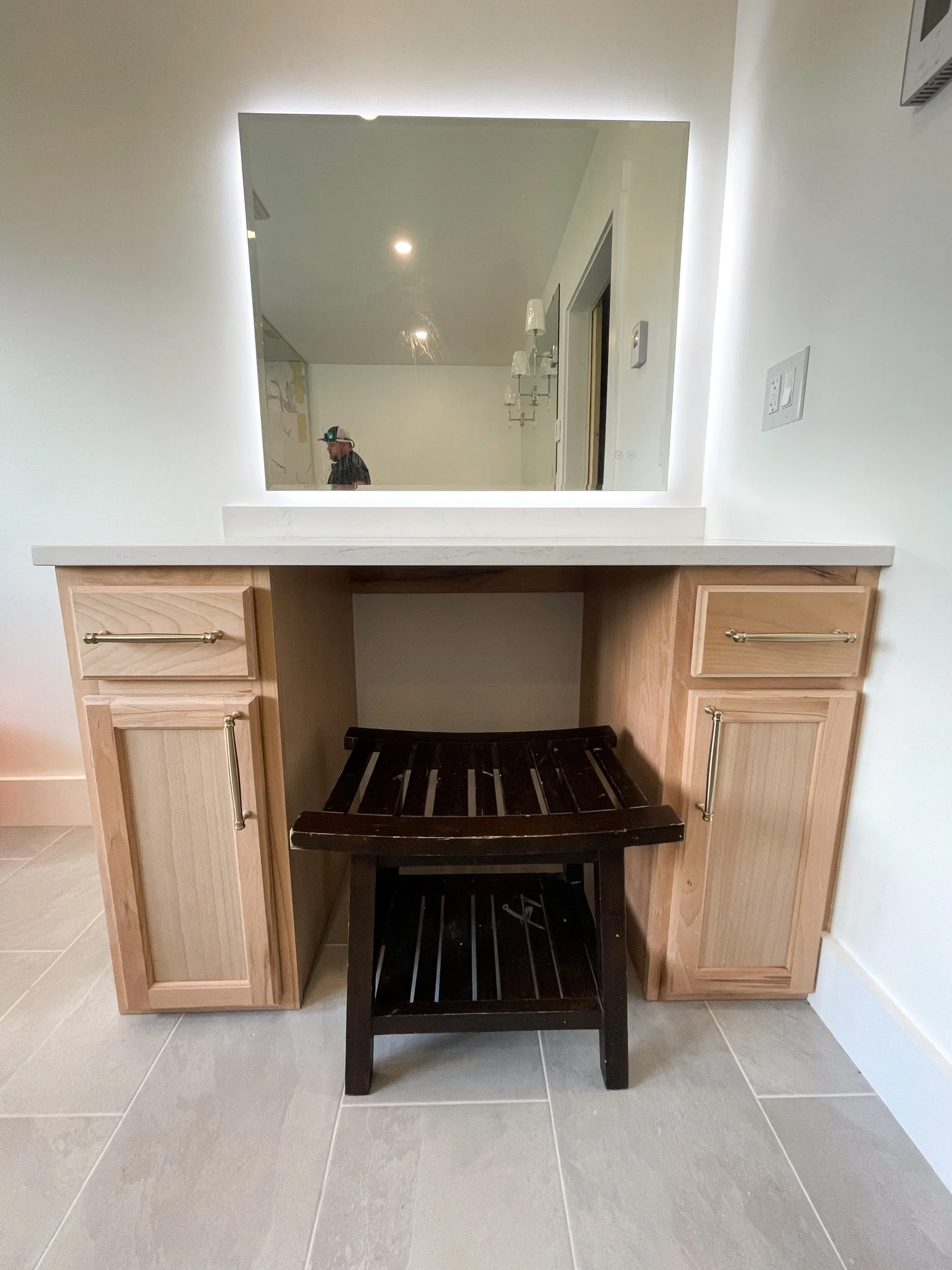 Bathroom vanity with a large mirror, wooden cabinets, and a dark wooden stool in front, with a person visible in the mirror reflection.