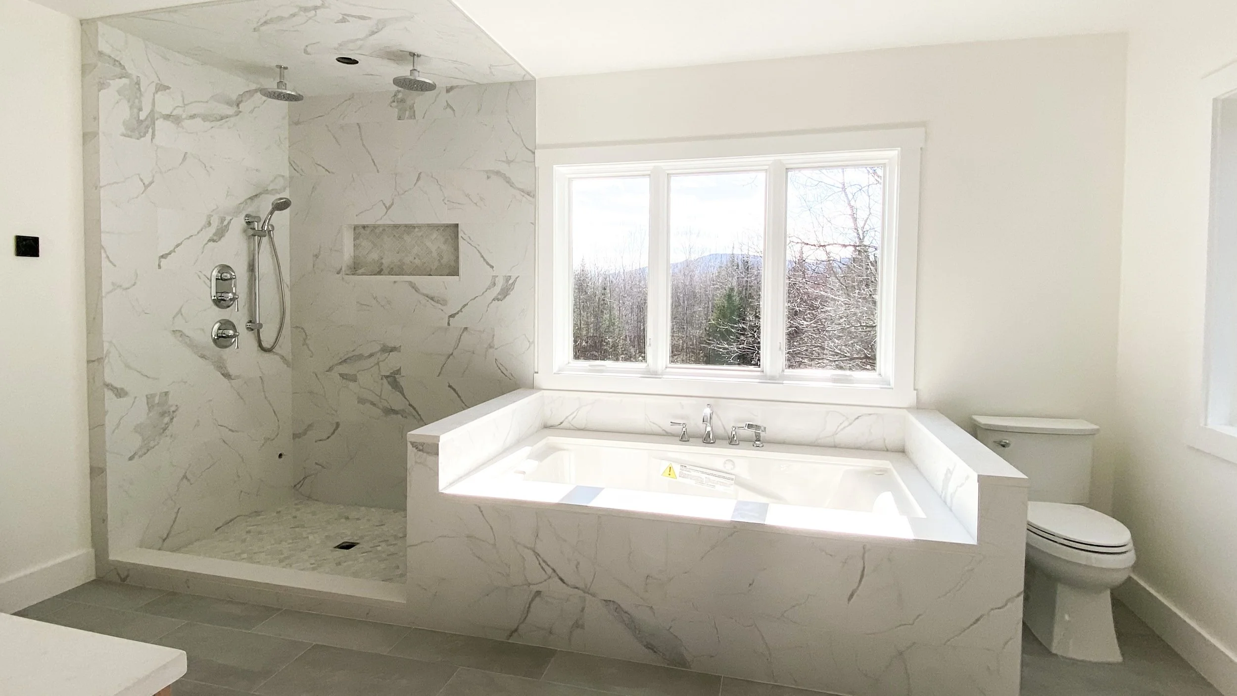 Modern bathroom with a large bathtub in front of a window, marble walls and a separate walk-in shower with showerheads, and a toilet to the right.
