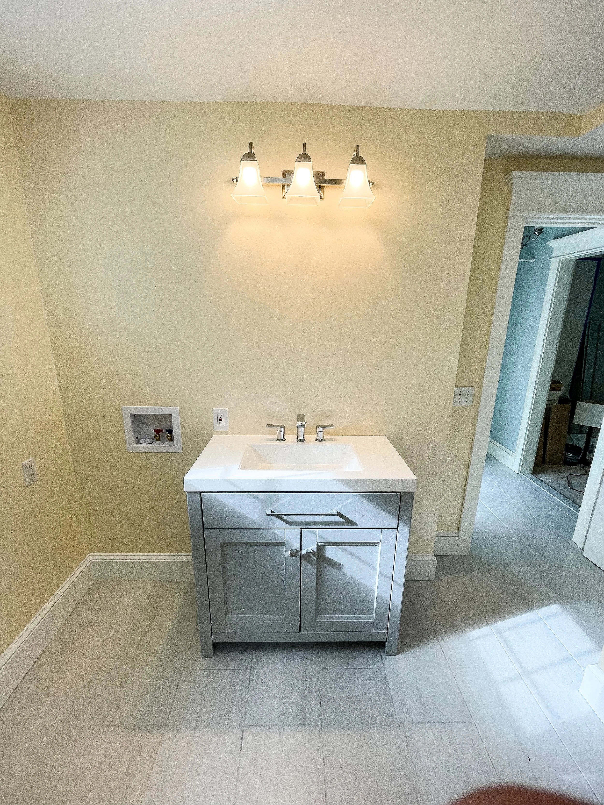 A small bathroom vanity with a white sink and a gray cabinet, illuminated by a three-light fixture above, with an empty plumbing connection area on the wall to the left.