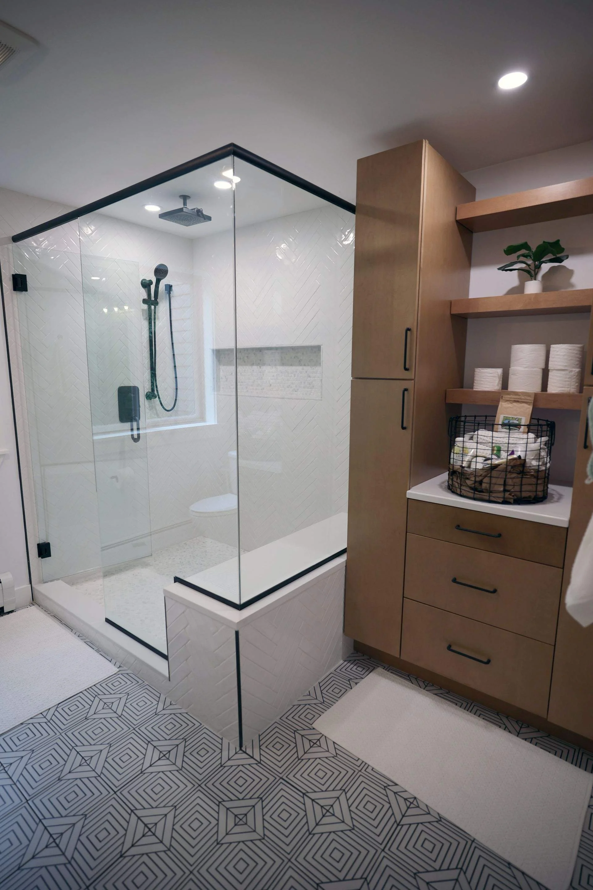 Modern bathroom with glass-enclosed shower, patterned floor tiles, and wooden shelving unit with toilet paper rolls and potted plant.
