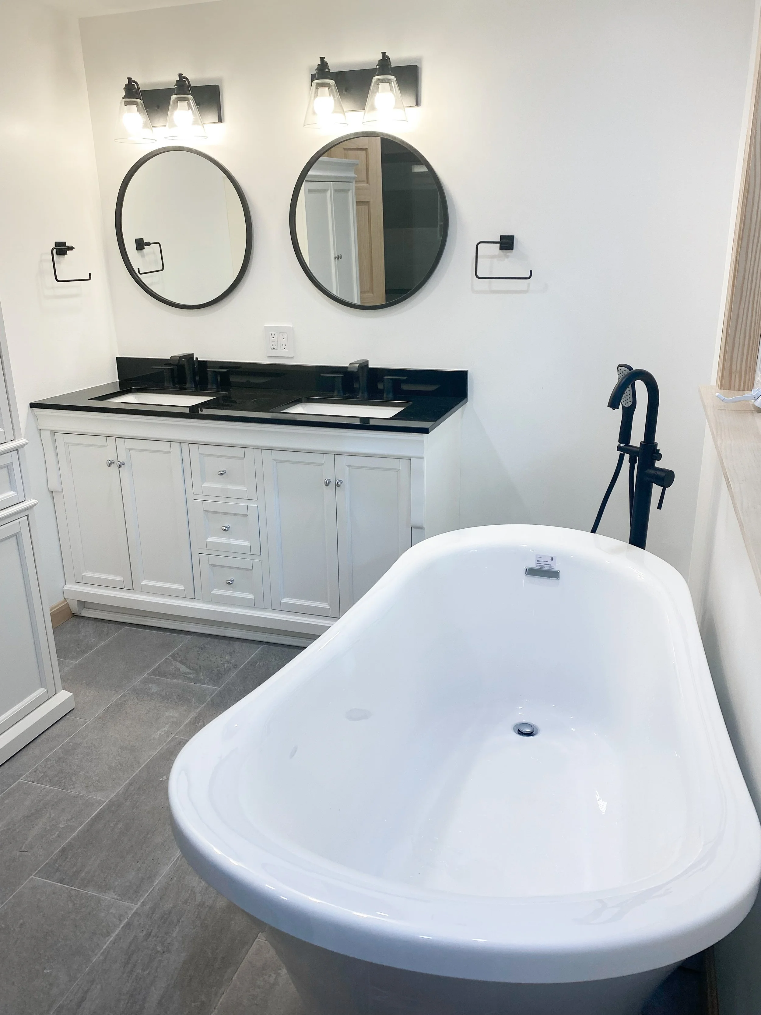 A bathroom featuring a large white soaking tub, a double vanity with black countertops, two circular mirrors, modern light fixtures above the mirrors, and gray tiled flooring.