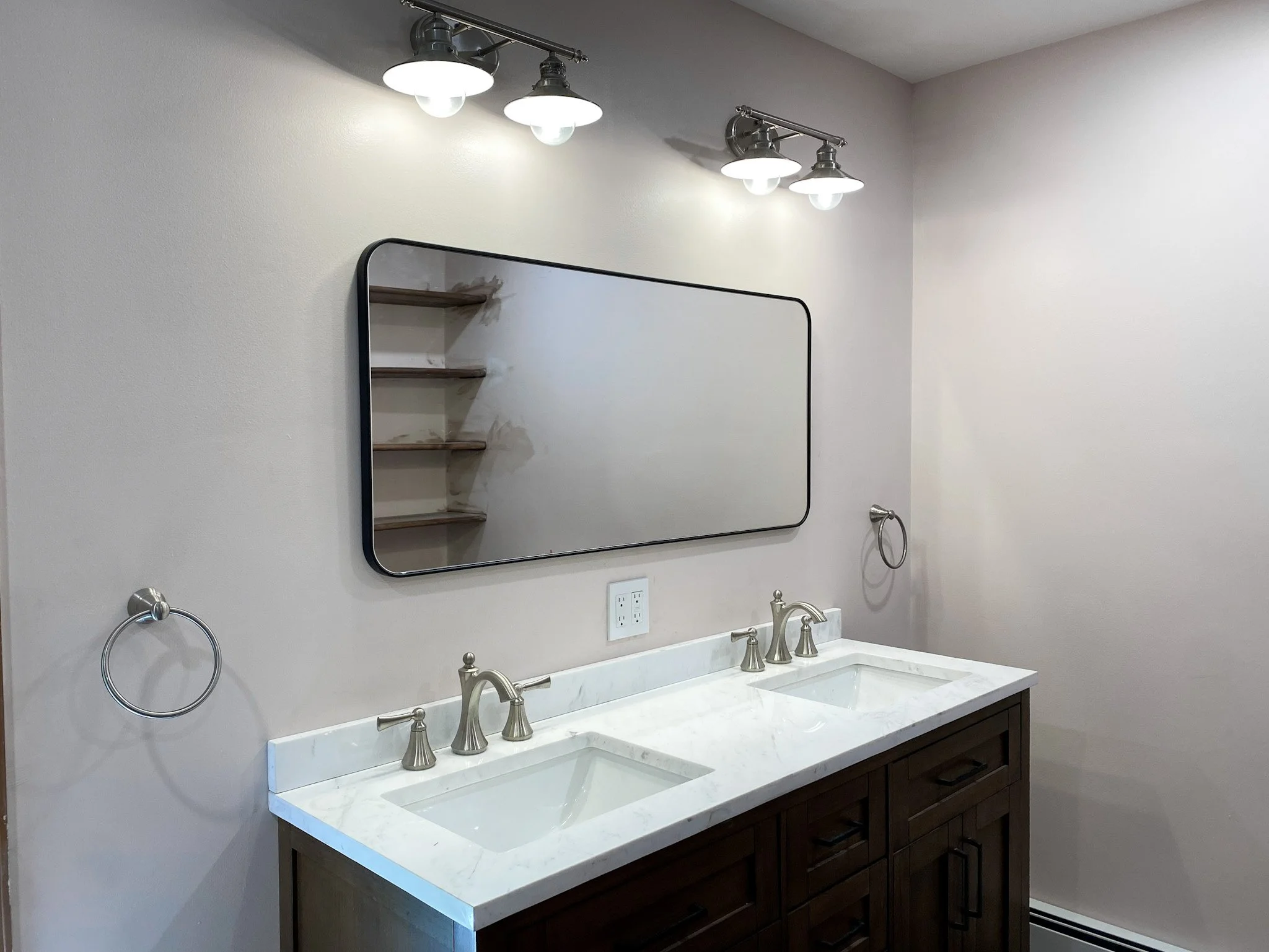 Bathroom vanity with double sinks, marble countertop, and a large rectangular mirror. Mounted above are two light fixtures with three bulbs each. There are two towel rings on either side of the mirror.