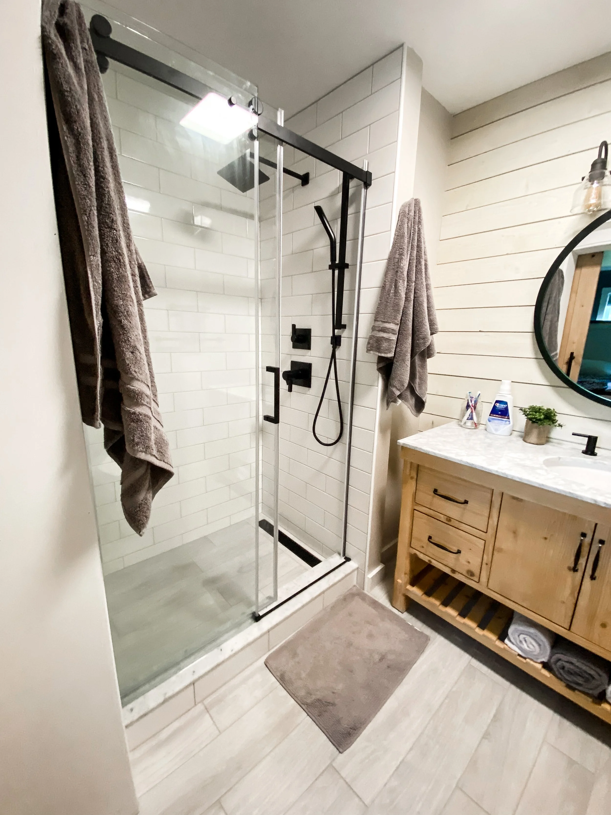 Bathroom with a glass-enclosed shower, beige tiles, a wooden vanity with a marble countertop, and towels hanging on the walls.