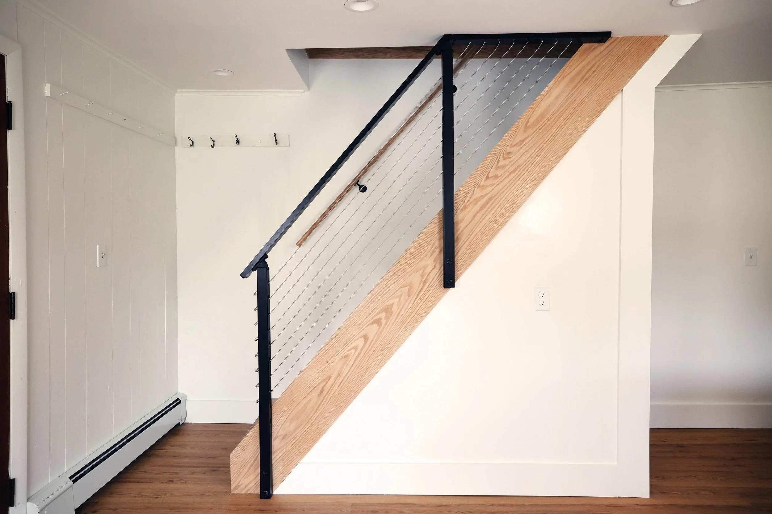 Interior view of a staircase with a wooden rail and metal cables, against white walls and a hardwood floor.