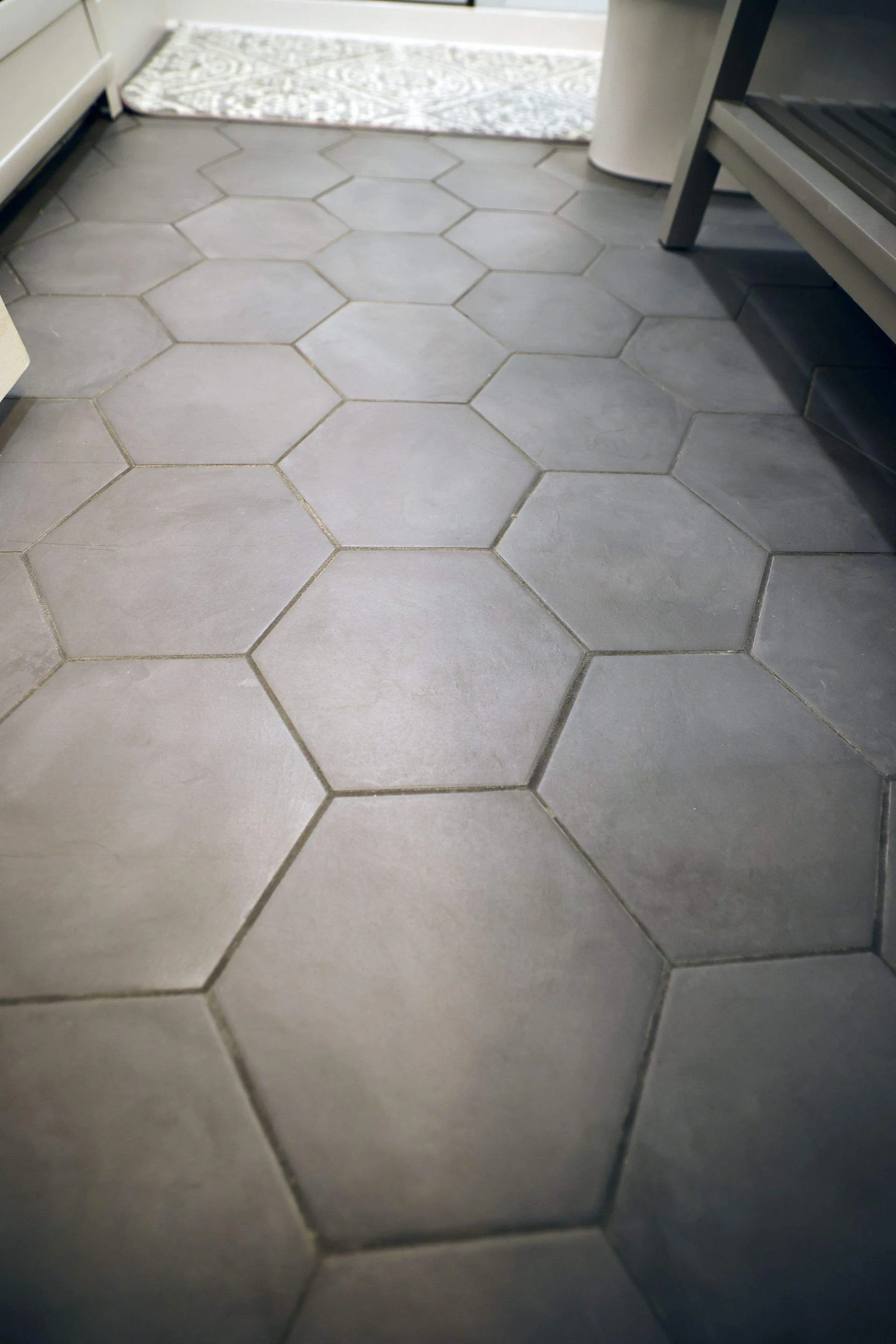 Close-up of hexagonal gray tiles on a kitchen floor, with part of a rug and kitchen cabinets visible.