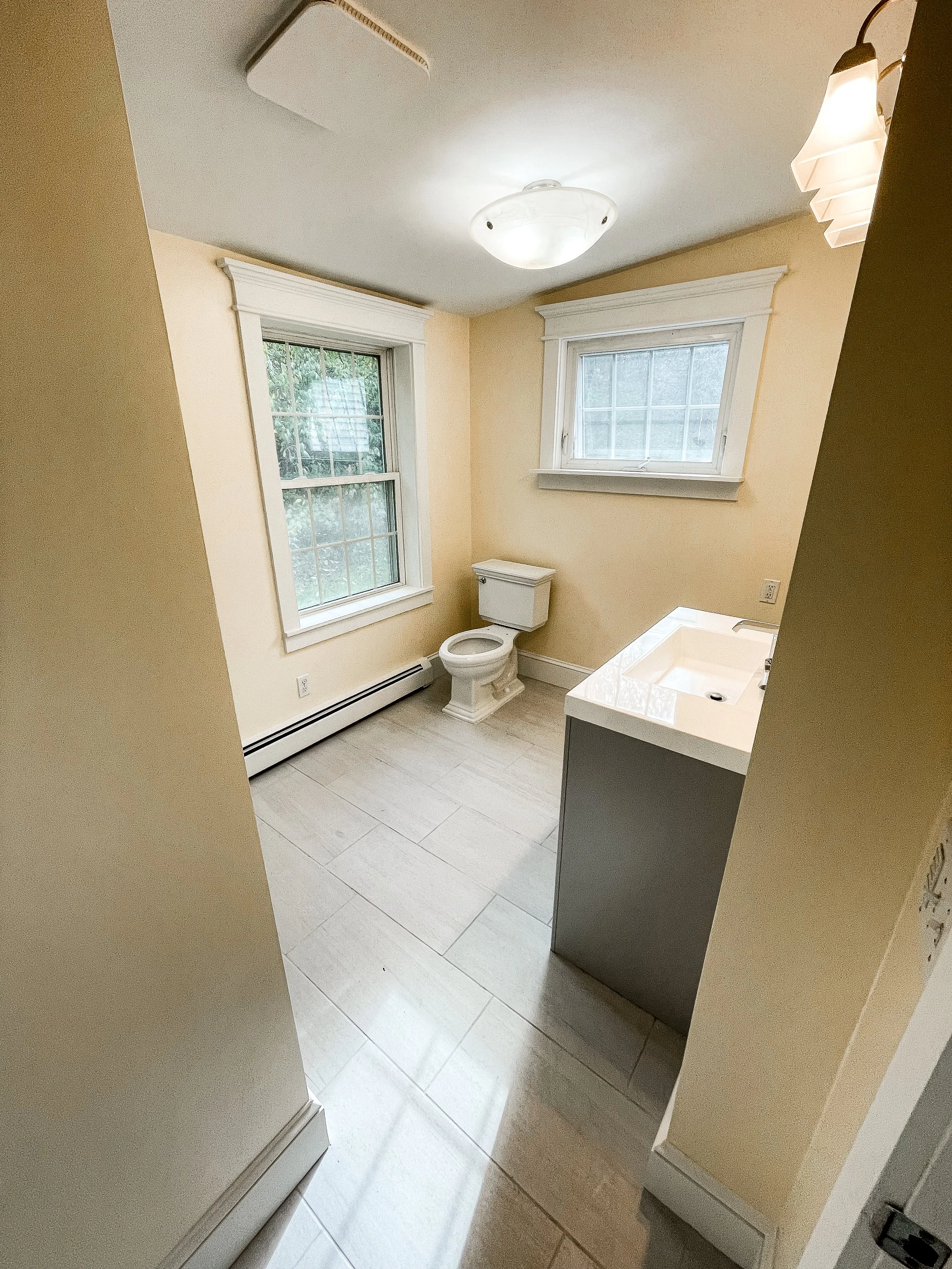 A small bathroom with beige walls, white trim, two windows, a toilet, a sink with a vanity, ceiling light, and tile floor.