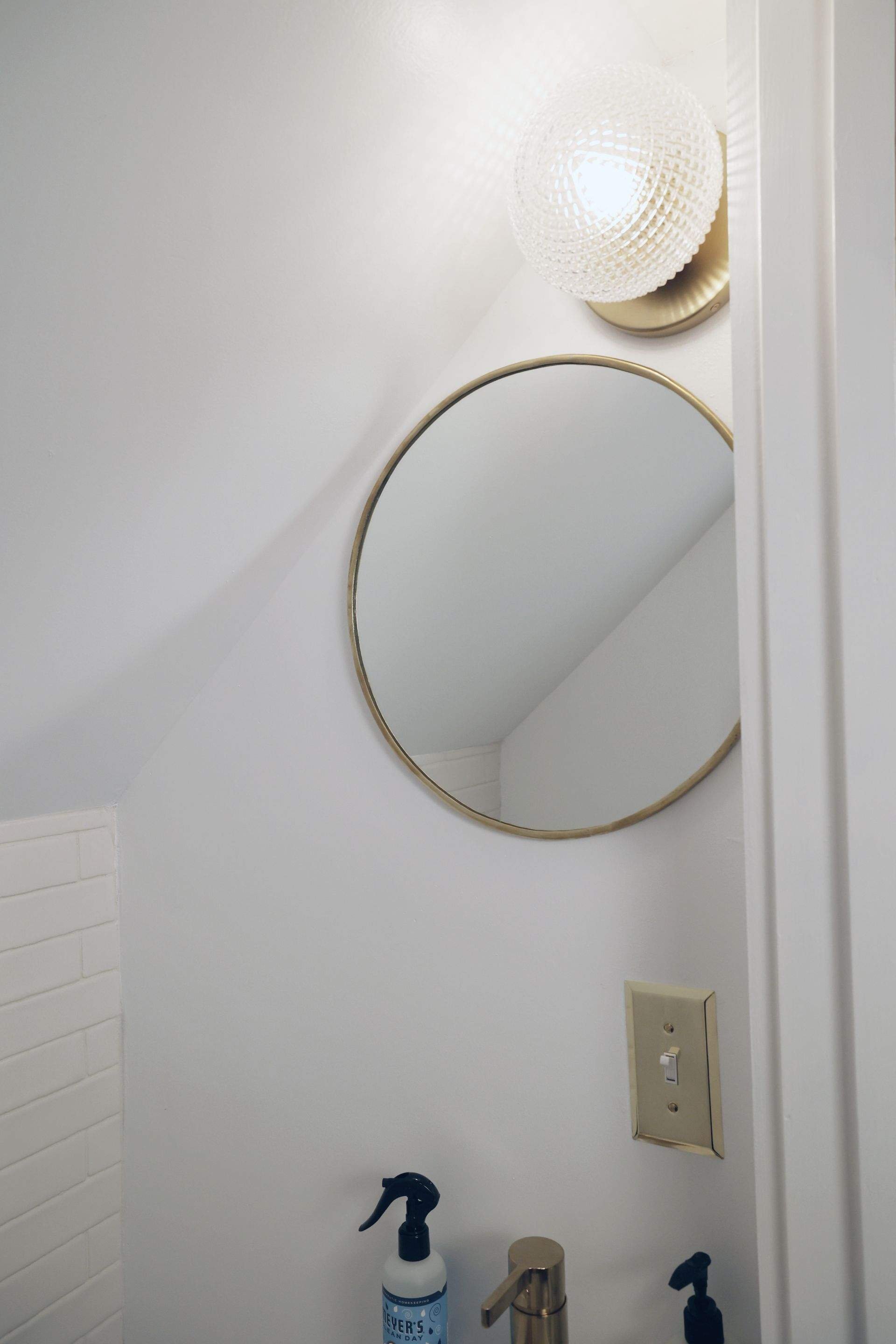 A bathroom wall with a round mirror, a light fixture, a light switch, and a sink with soap bottles.