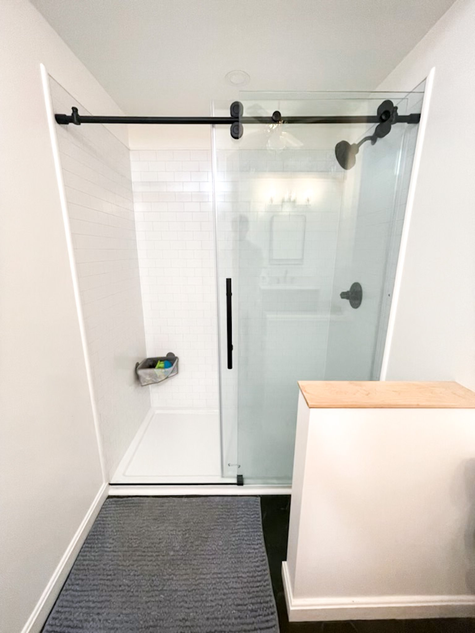Bathroom shower with glass door, black sliding door hardware, white tiled walls, rain showerhead, small storage basket, gray bath mat, and white walls.