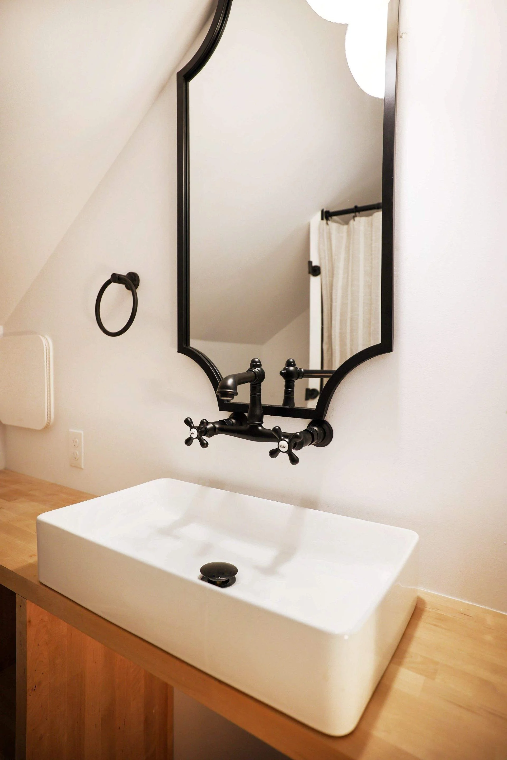 Bathroom sink with a white rectangular ceramic basin, black vintage-style faucet with cross handles, mounted on a wooden countertop, black-framed mirror with a curved edge, black towel ring, and a striped shower curtain in the background.
