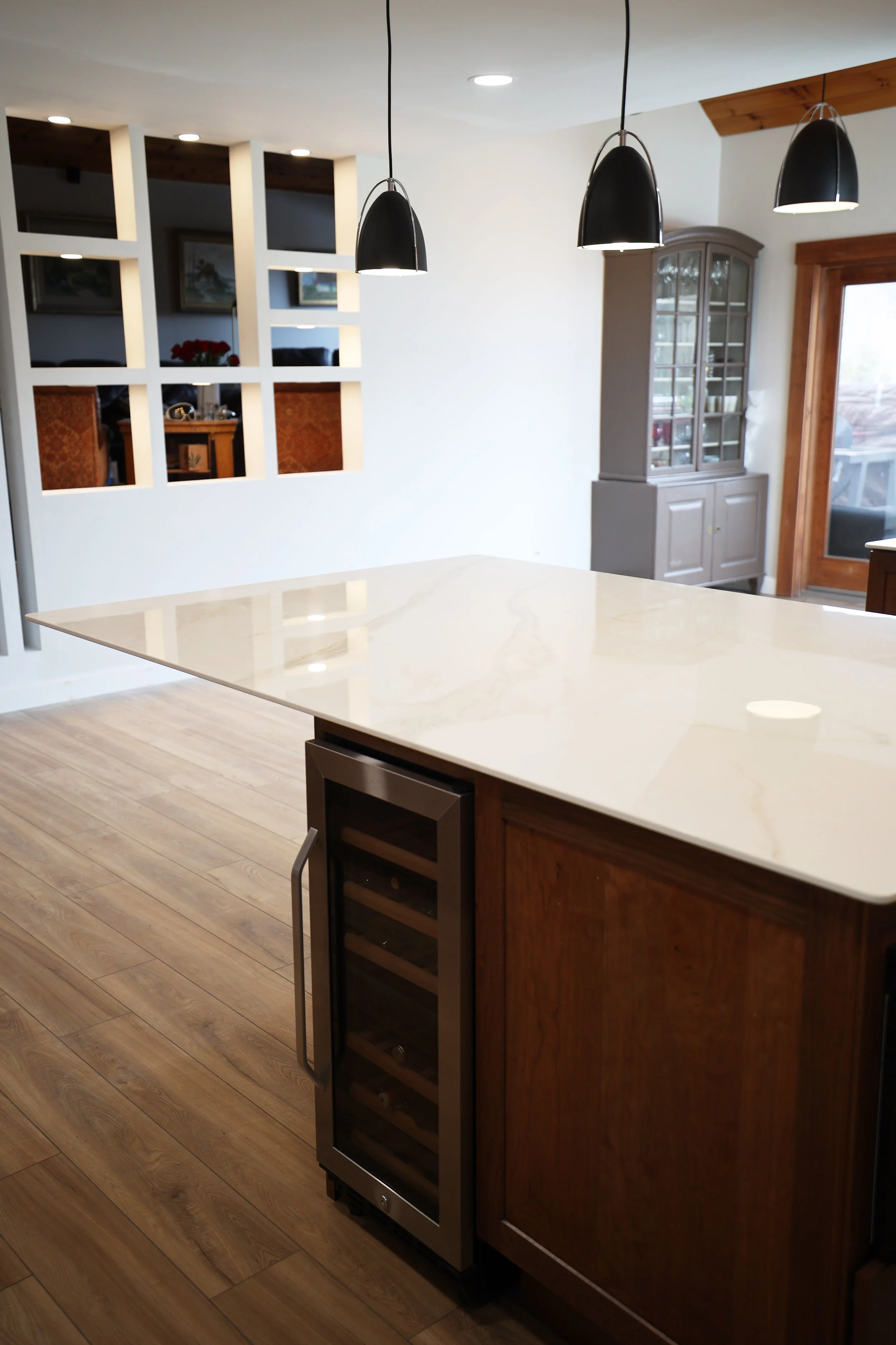 Kitchen area featuring a white marble countertop on a wooden island, black pendant lights hanging above, and a built-in wine cooler at the end of the island. In the background, there is a gray cupboard with glass doors and a wooden framed sliding door leading outside.
