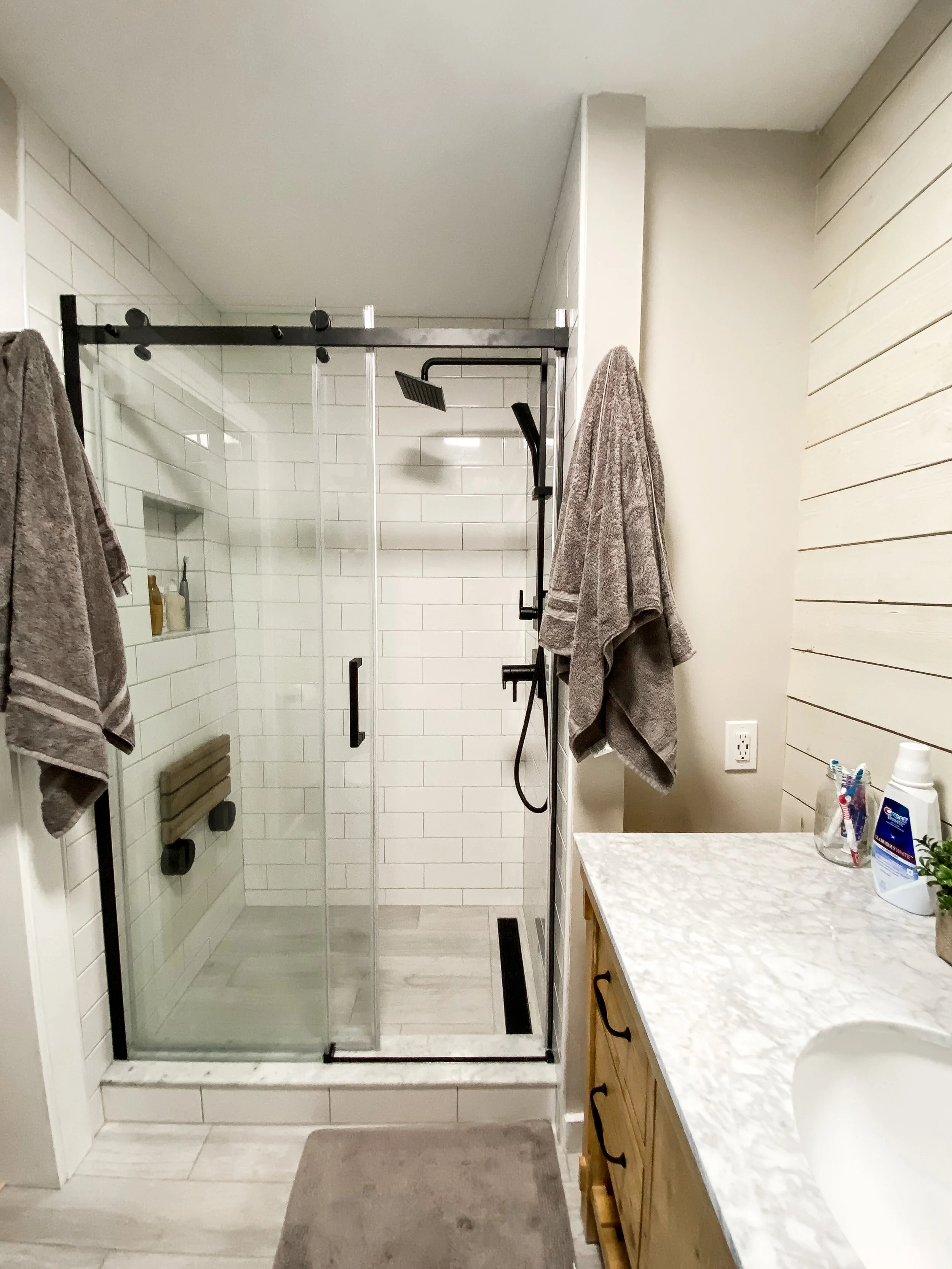 Modern bathroom with a glass shower enclosure, white subway tile walls, a beige wooden vanity with a marble countertop, and towels hanging on the walls.