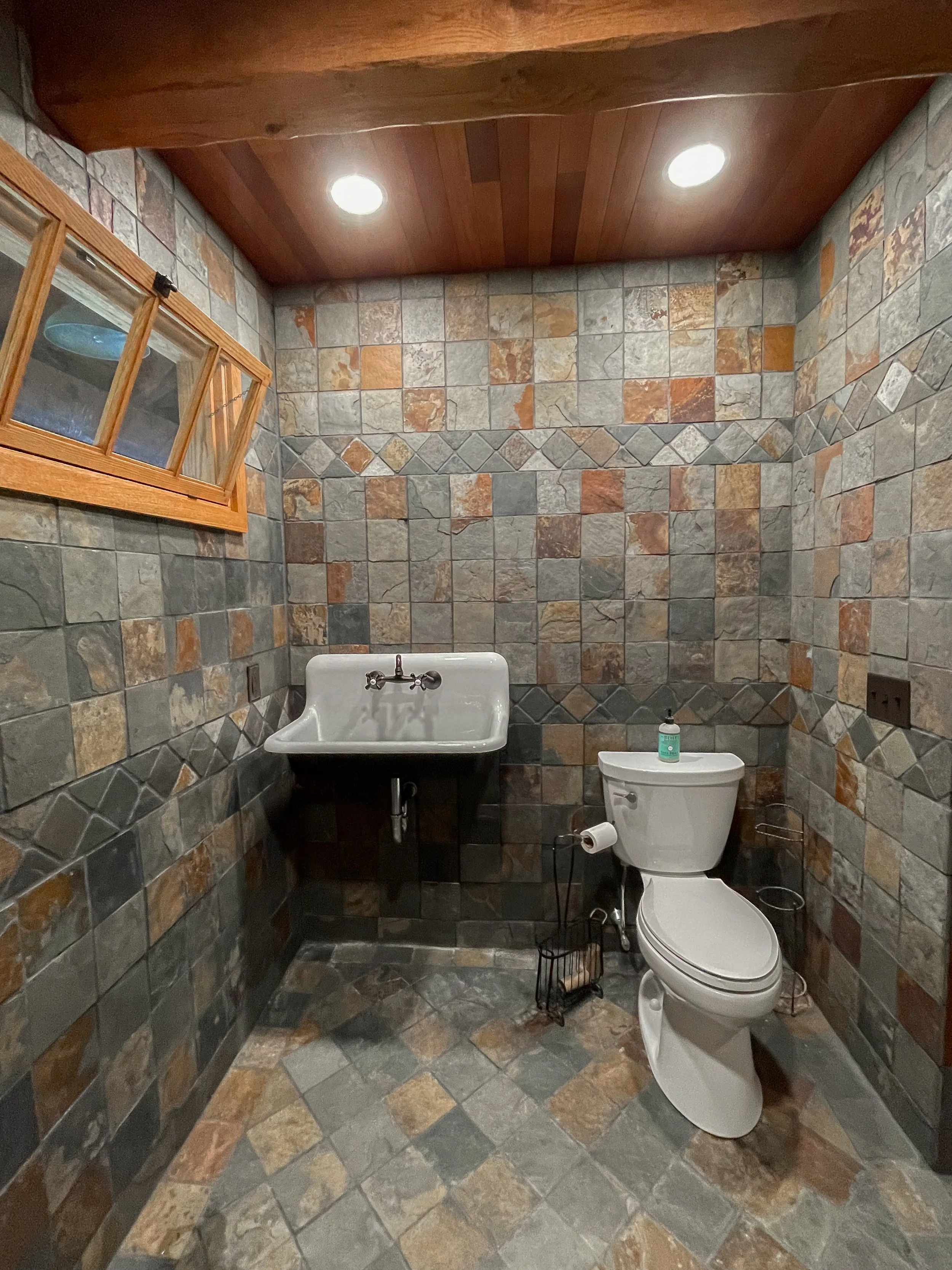 Bathroom with rustic stone tile walls and floor, a white toilet, a wall-mounted white utility sink, and a small window with wooden trim, ceiling lighting