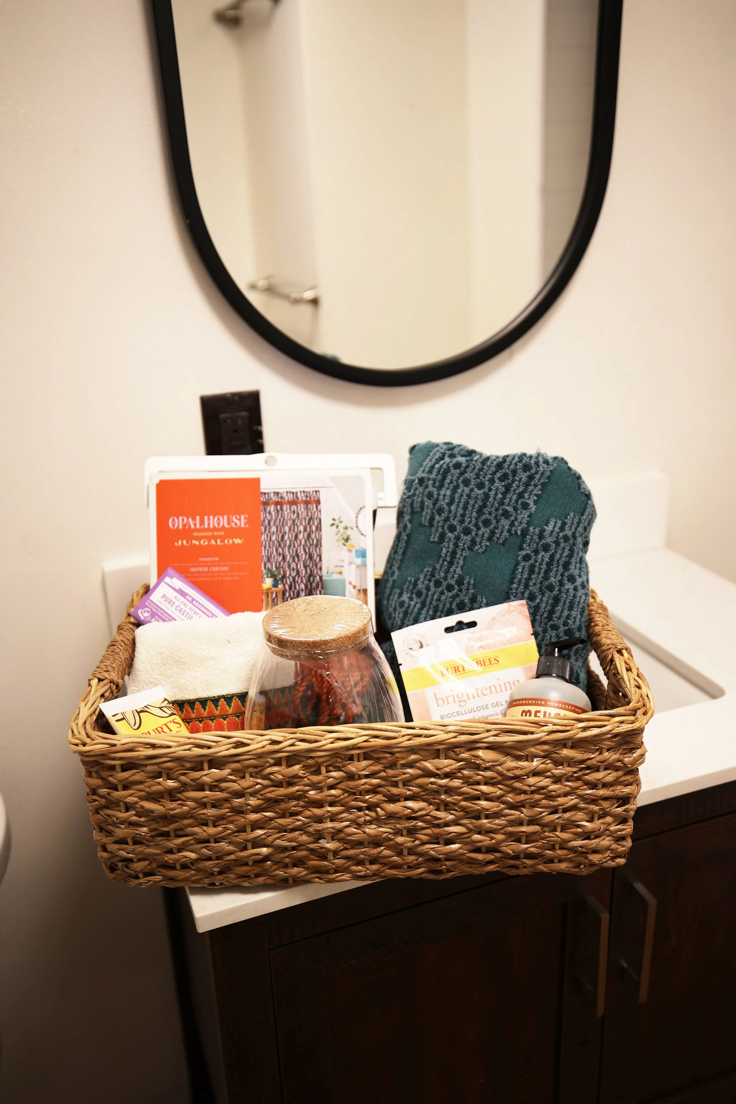 A wicker basket filled with various personal care and household items, placed on a white countertop in front of a wall mirror.