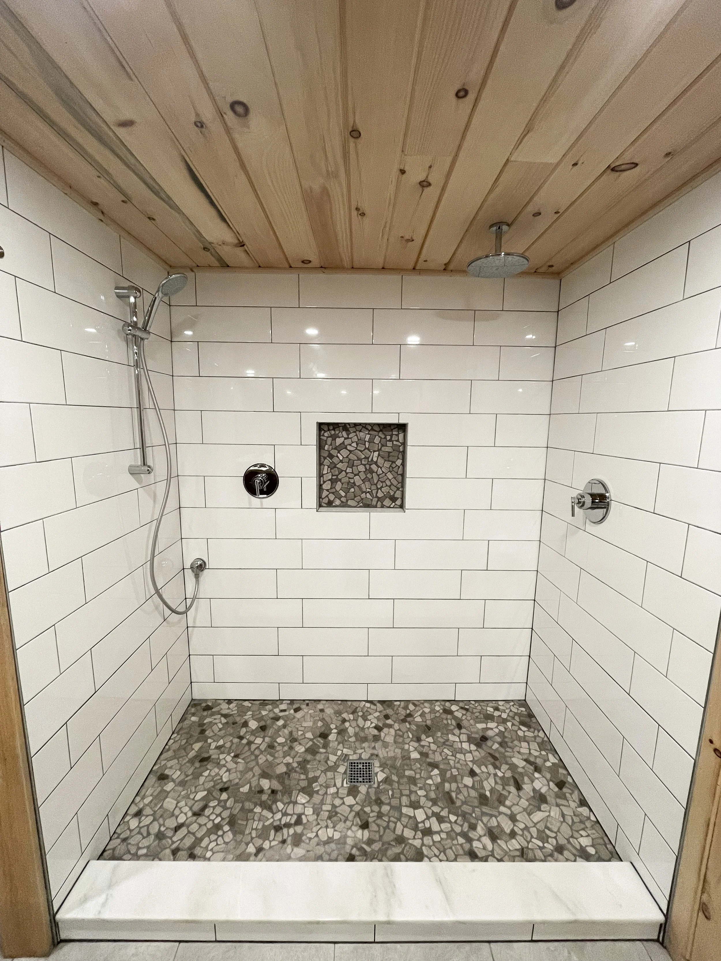 A walk-in shower with white subway tile walls, a pebble tile floor, a built-in niche with pebbles, a rainfall showerhead, a handheld showerhead, and chrome fixtures, framed by a wooden ceiling and trim.