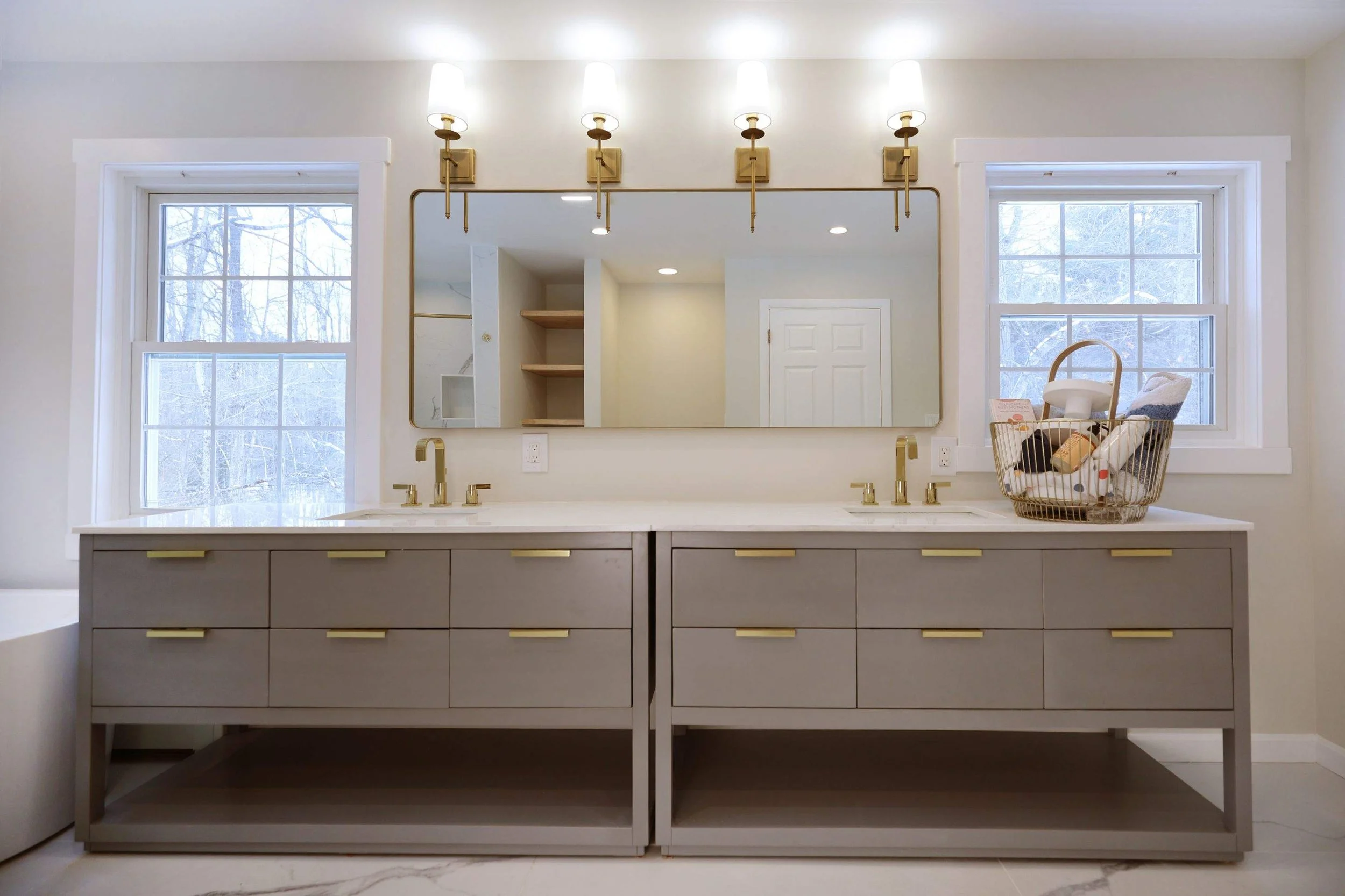A modern bathroom vanity with a large mirror above it, two sinks with gold fixtures, and a basket of toiletries on the right side near a window. The room has white walls and two windows showing snowy trees outside.