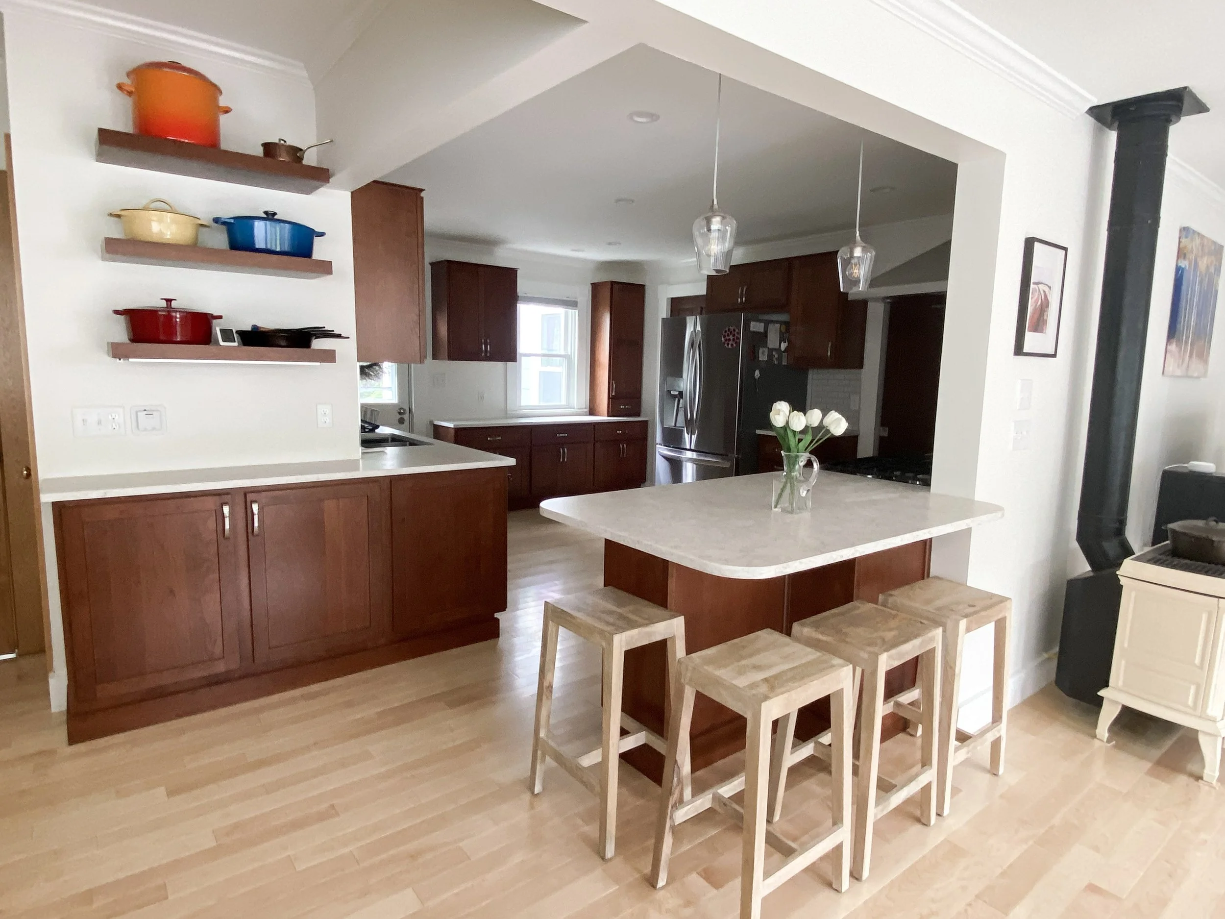 Kitchen with wooden cabinets, a kitchen island with four wooden stools, and open shelves with colorful pots and pans. There is a stainless steel refrigerator, pendant lighting, and a vase with white roses on the island. The floor is light wood.