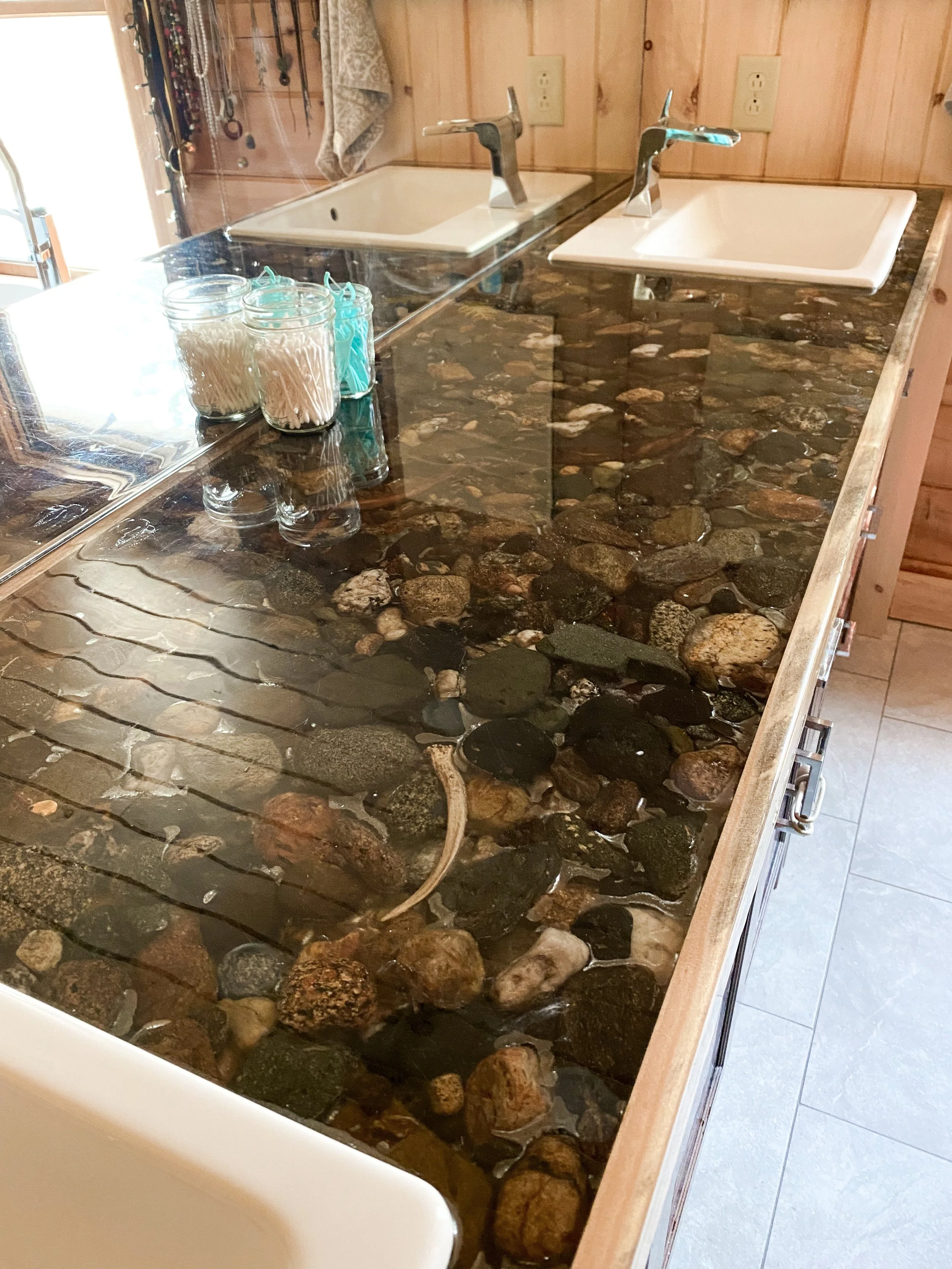 A kitchen countertop made of resin with embedded river rocks, featuring two white sinks with faucets, jars of cotton swabs and pads, and a wooden wall with electrical outlets.