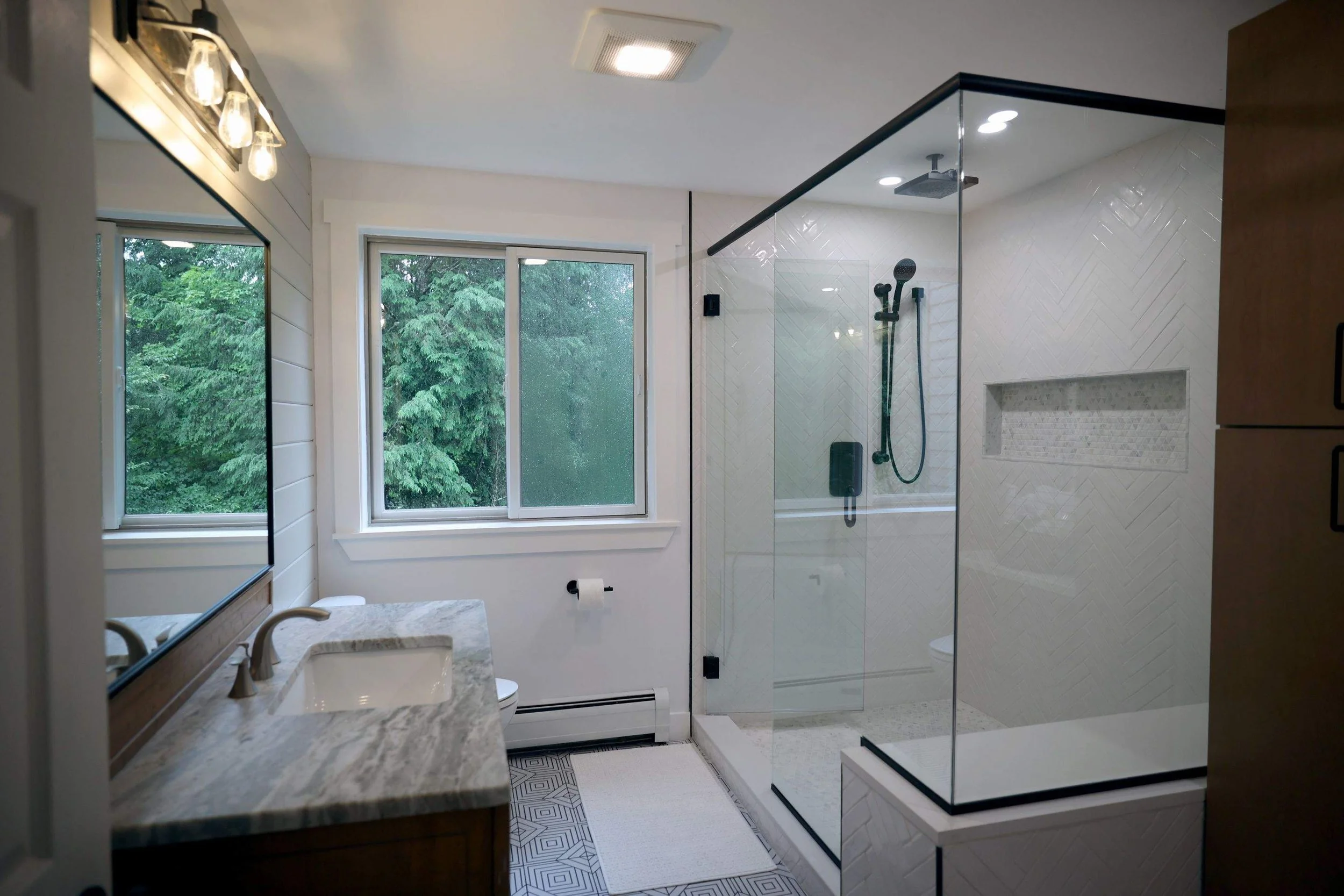 Bathroom with a large window showing green trees, a granite countertop sink, a mirror with light bulbs, a glass-enclosed shower with tiled walls, and a toilet.