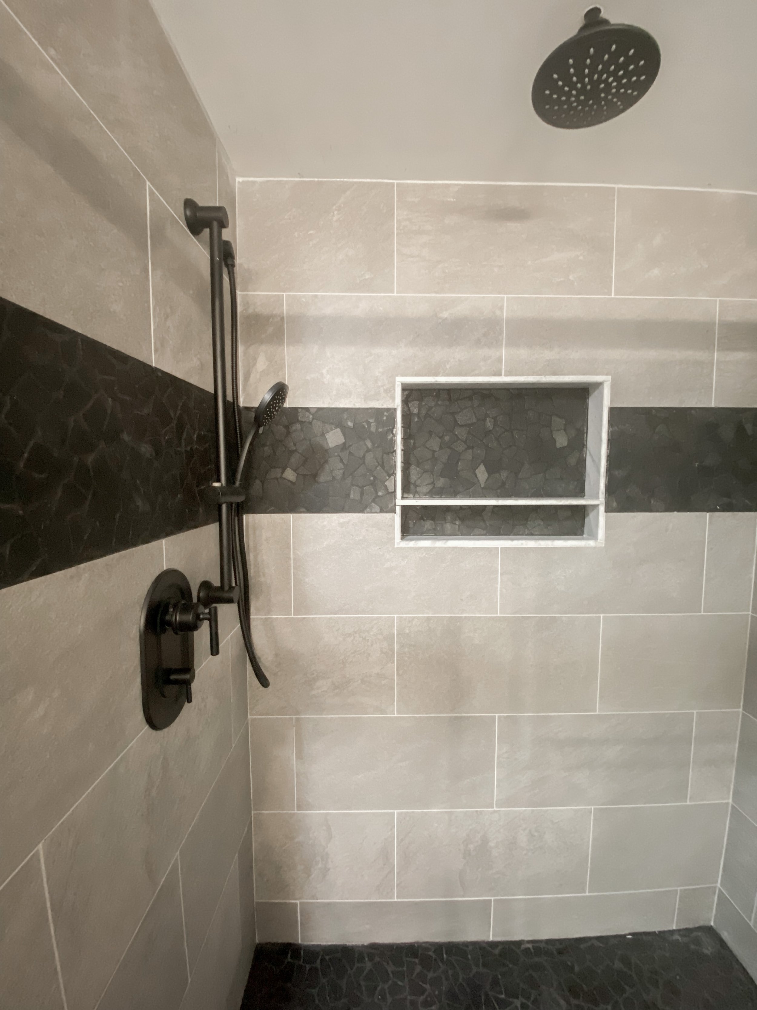 Shower with beige and black tile walls, a black showerhead, a black handle, and a built-in rectangular shelf.