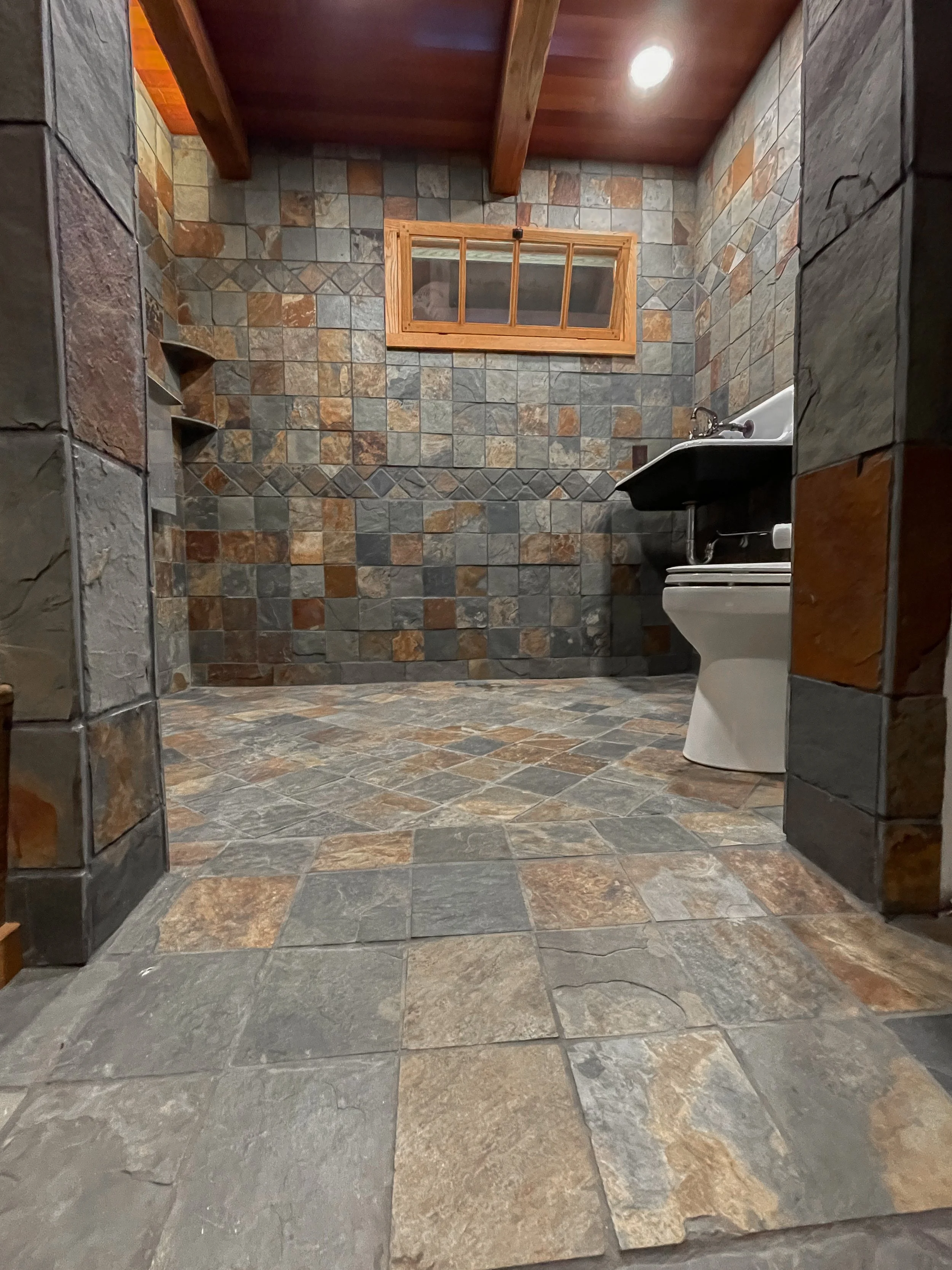 Bathroom with stone-tile walls and floor, wooden ceiling, small window, and black sink with white toilet.