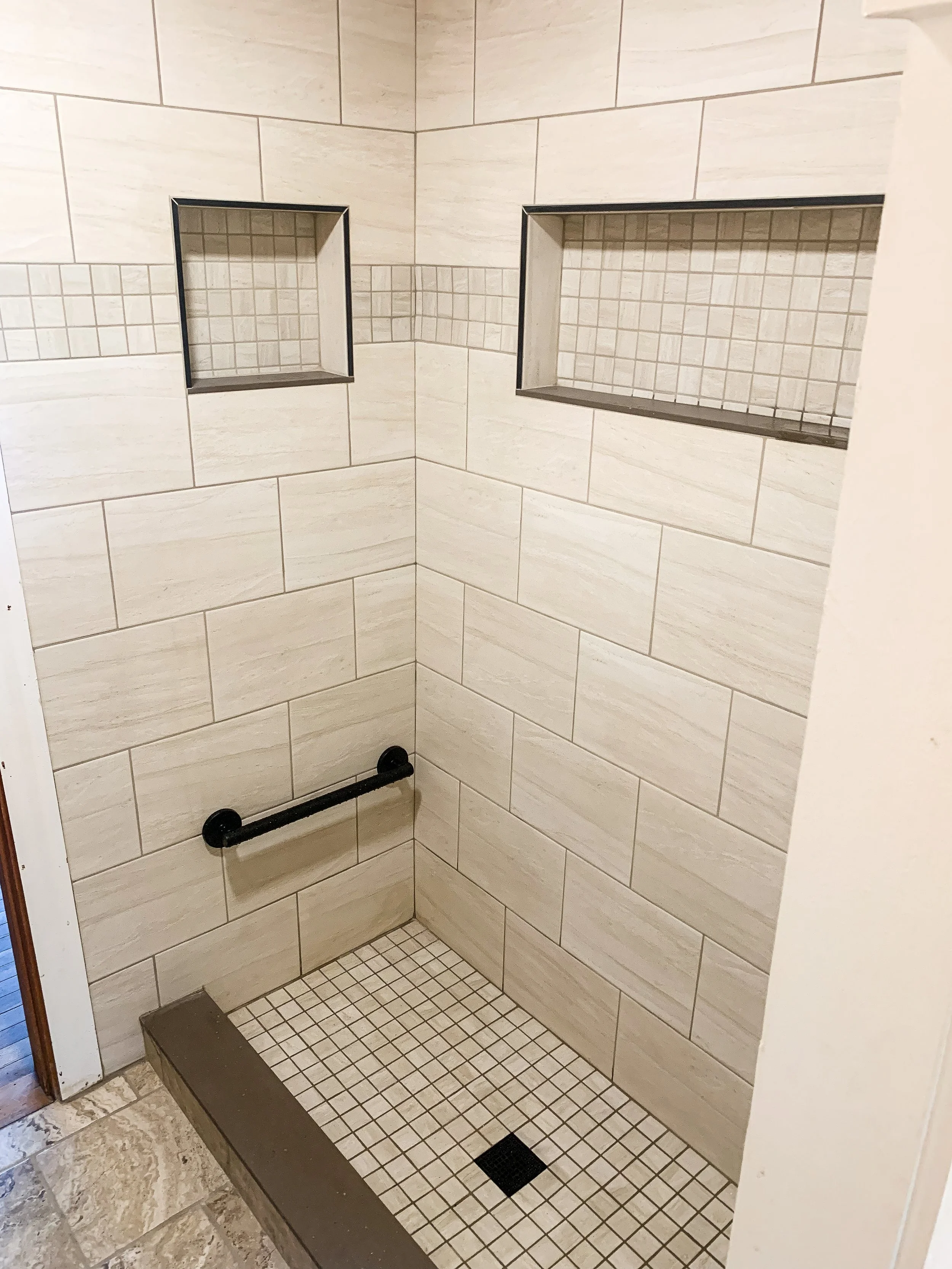 A walk-in shower with beige large tiles on the wall, two built-in shelves with small tiles, a black grab bar, and small beige and black tiles on the shower floor.