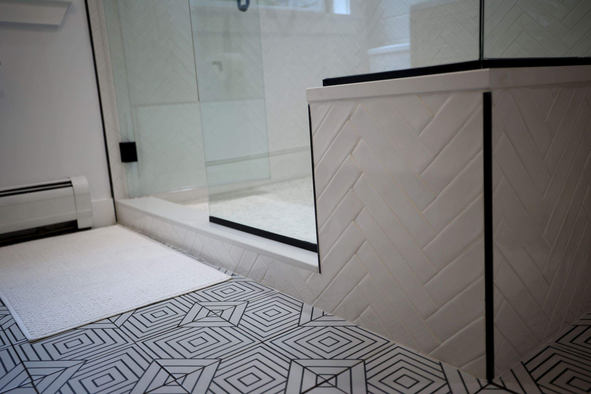 Close-up of a bathroom vanity with herringbone white tile, black grout, and a mirror, next to a tiled shower with a glass door and geometric floor pattern.