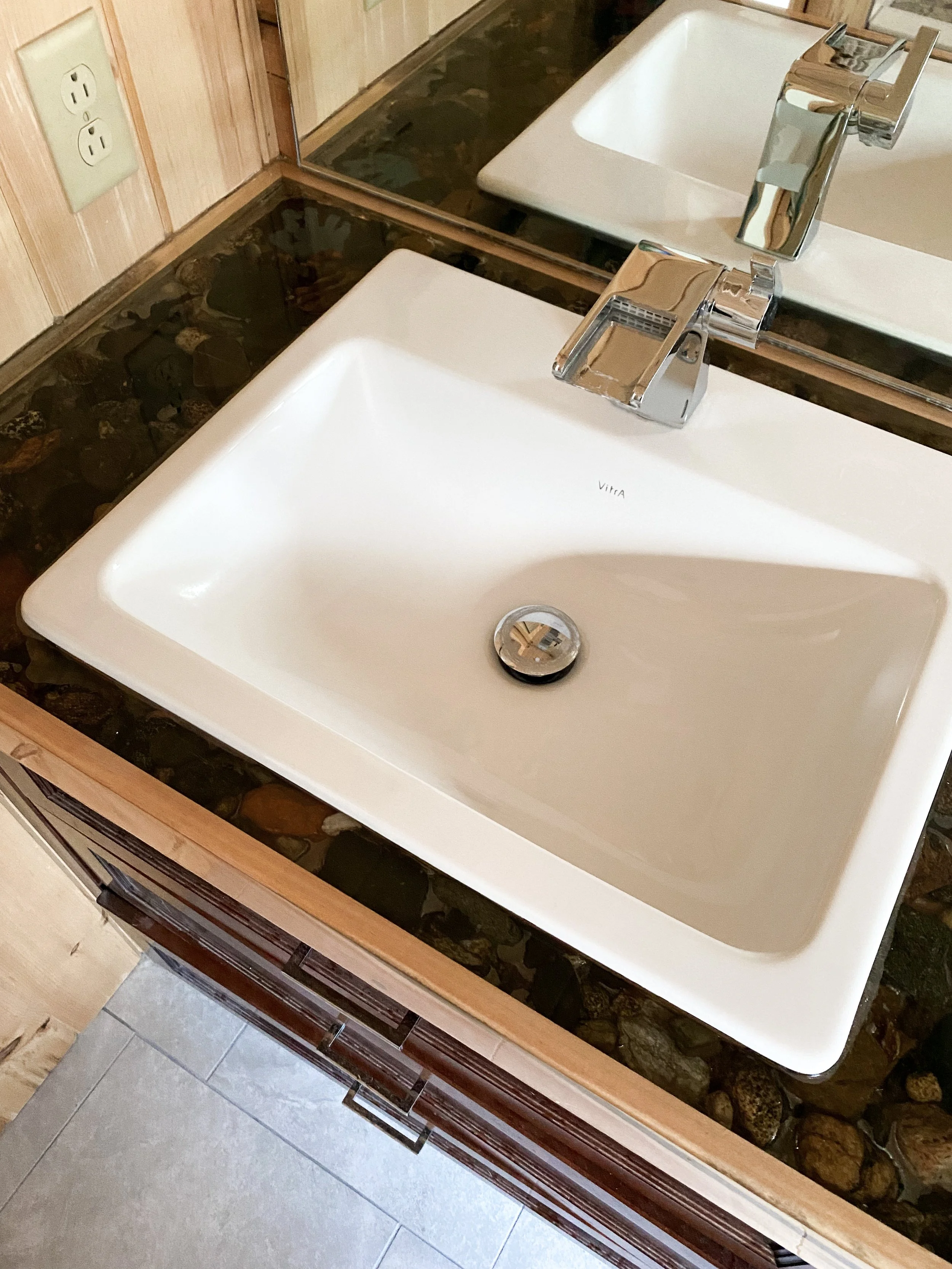 Bathroom sink with a modern chrome faucet, set in a countertop with decorative stones underneath and a large mirror behind, alongside a wall with electrical outlets.