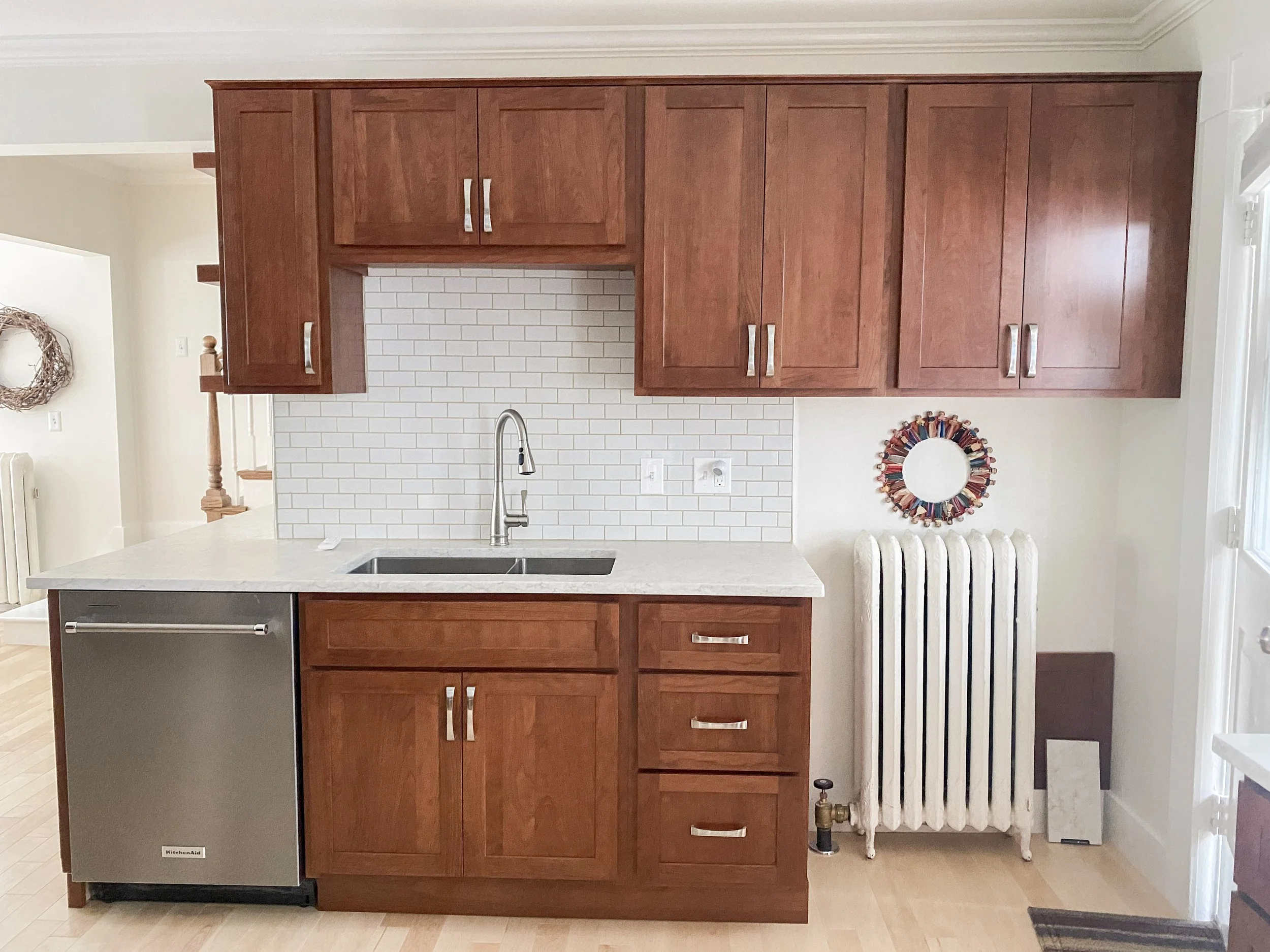 Kitchen with wooden cabinets, white backsplash, stainless steel sink, small fridge, and vintage radiator.