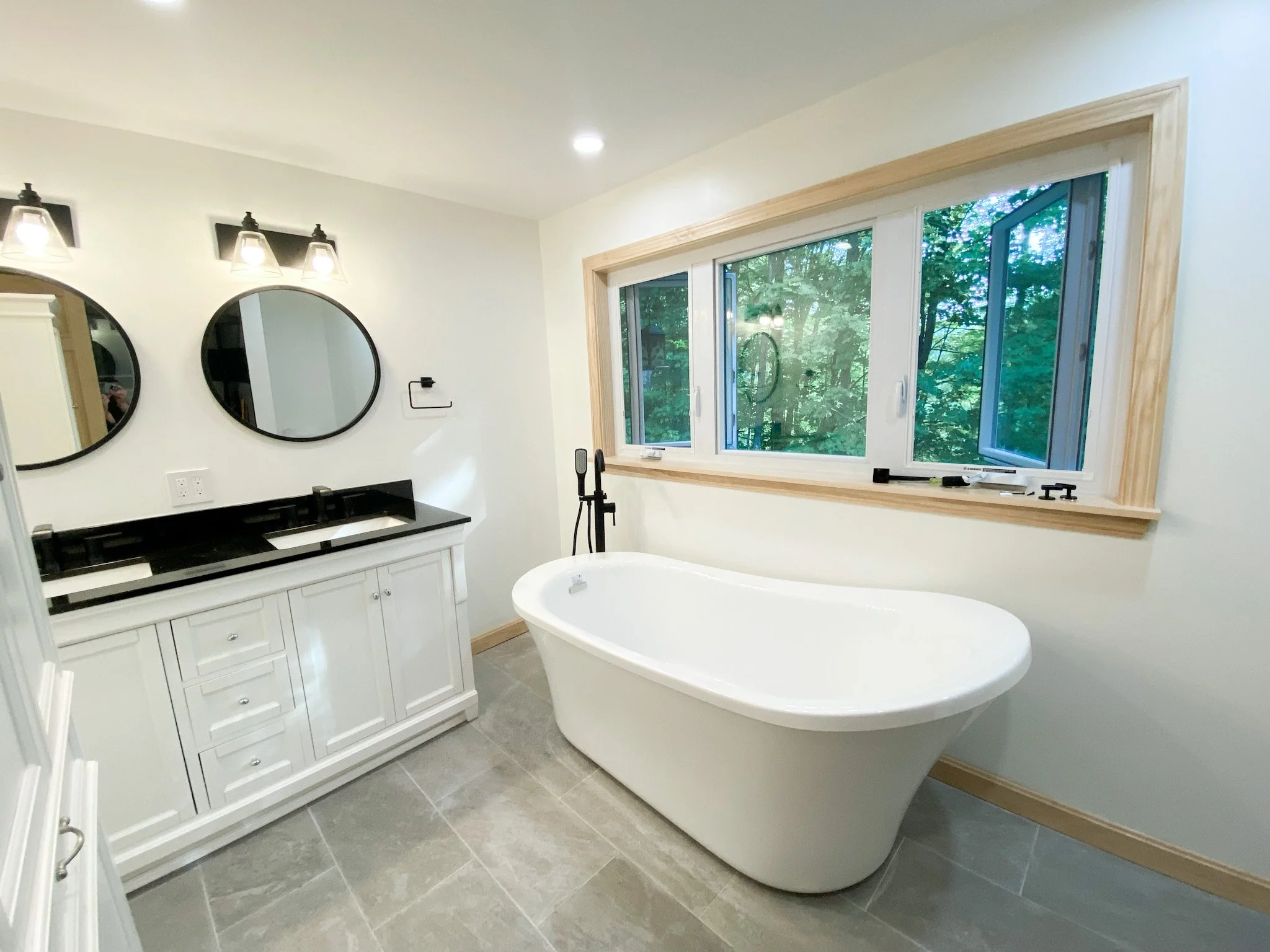 Bathroom with a freestanding bathtub, a white vanity with a black countertop, two round mirrors, and a large window overlooking greenery.