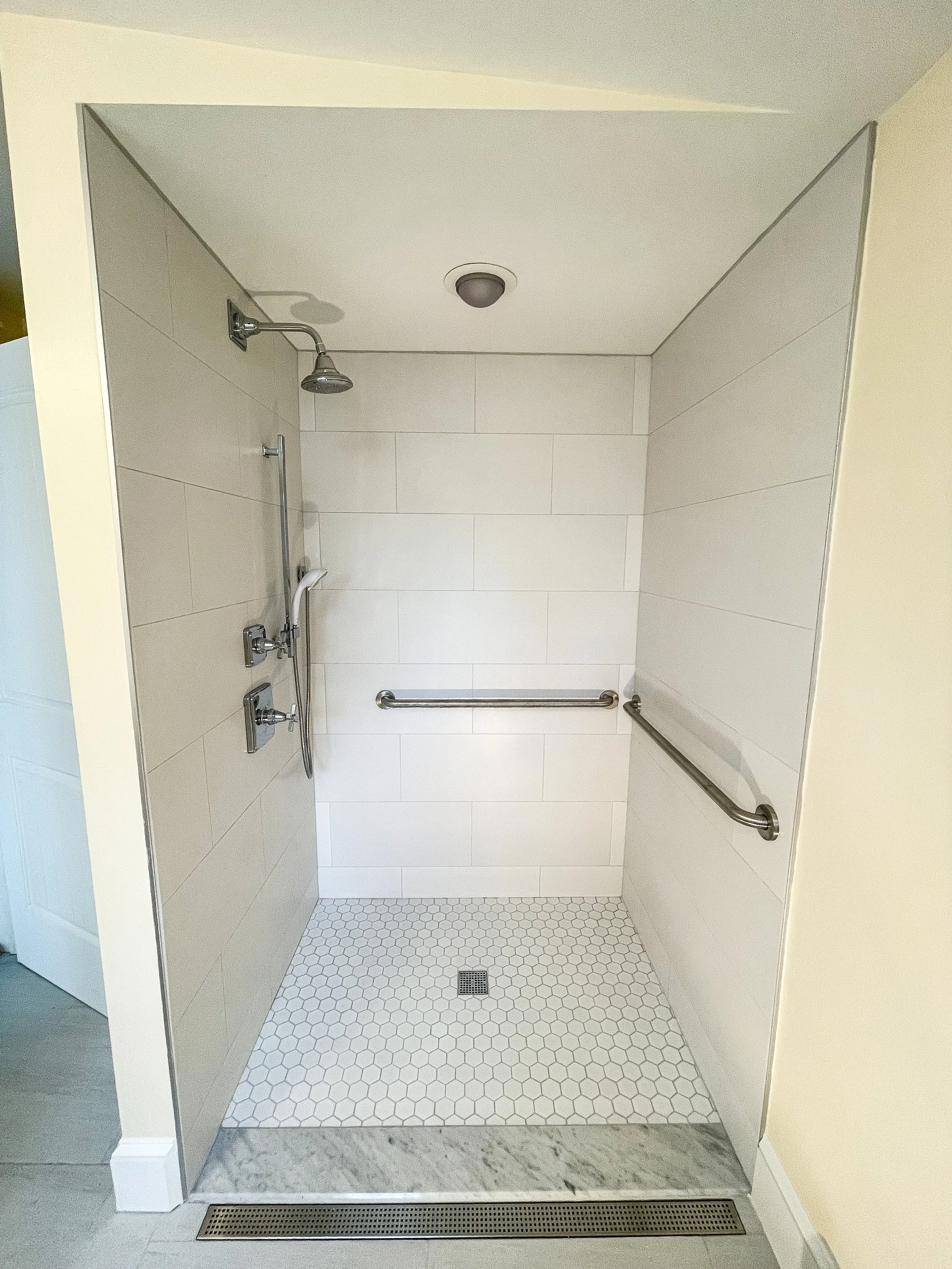 Accessible shower with a ceiling-mounted showerhead, handheld shower, grab bars on the walls, and a tiled floor with a drain, designed for disabled access.