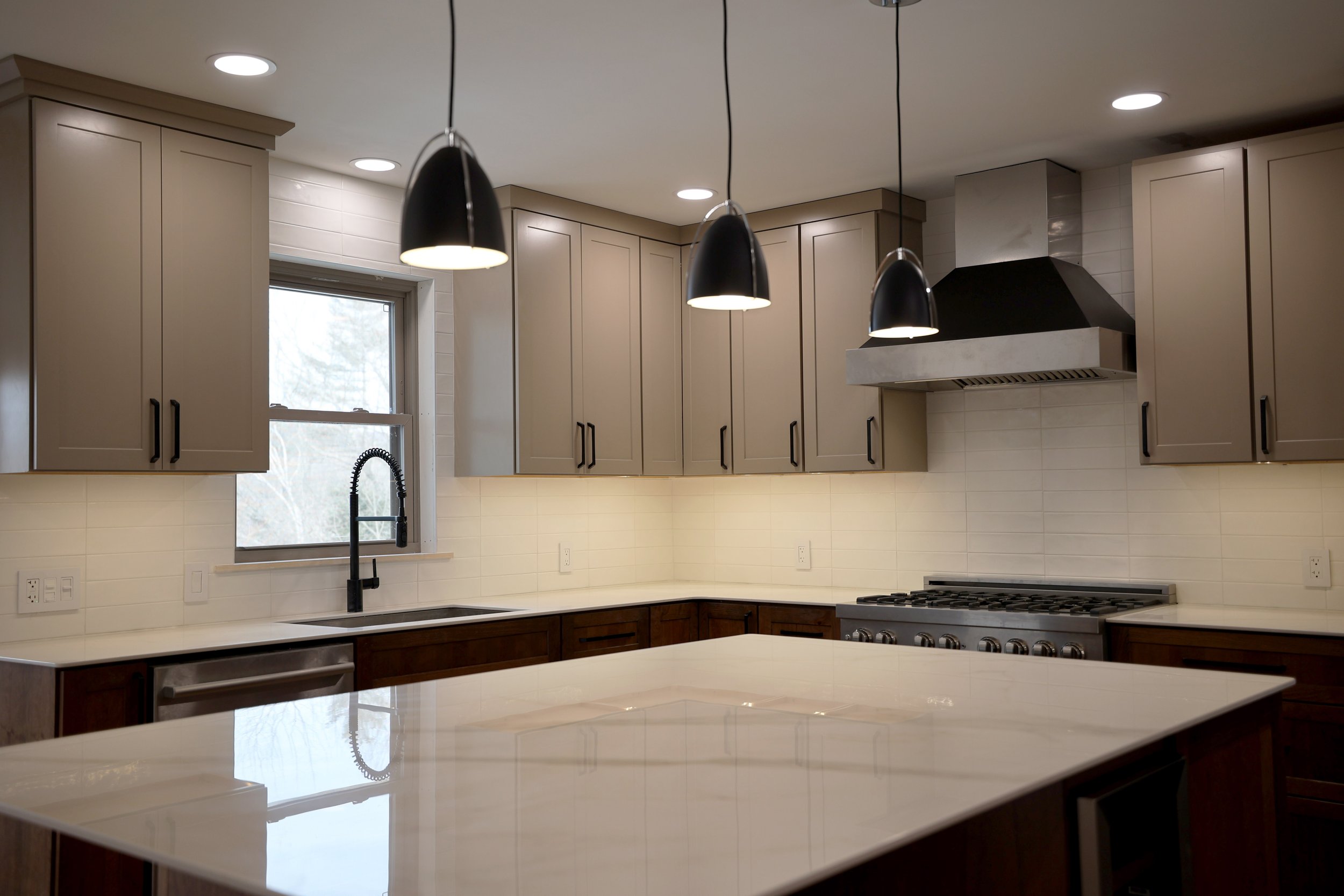 Modern kitchen with beige upper cabinets, dark lower cabinets, white countertop, four black pendant lights, stainless steel stove with matching range hood, white tiled backsplash, sink under a window, and natural light.