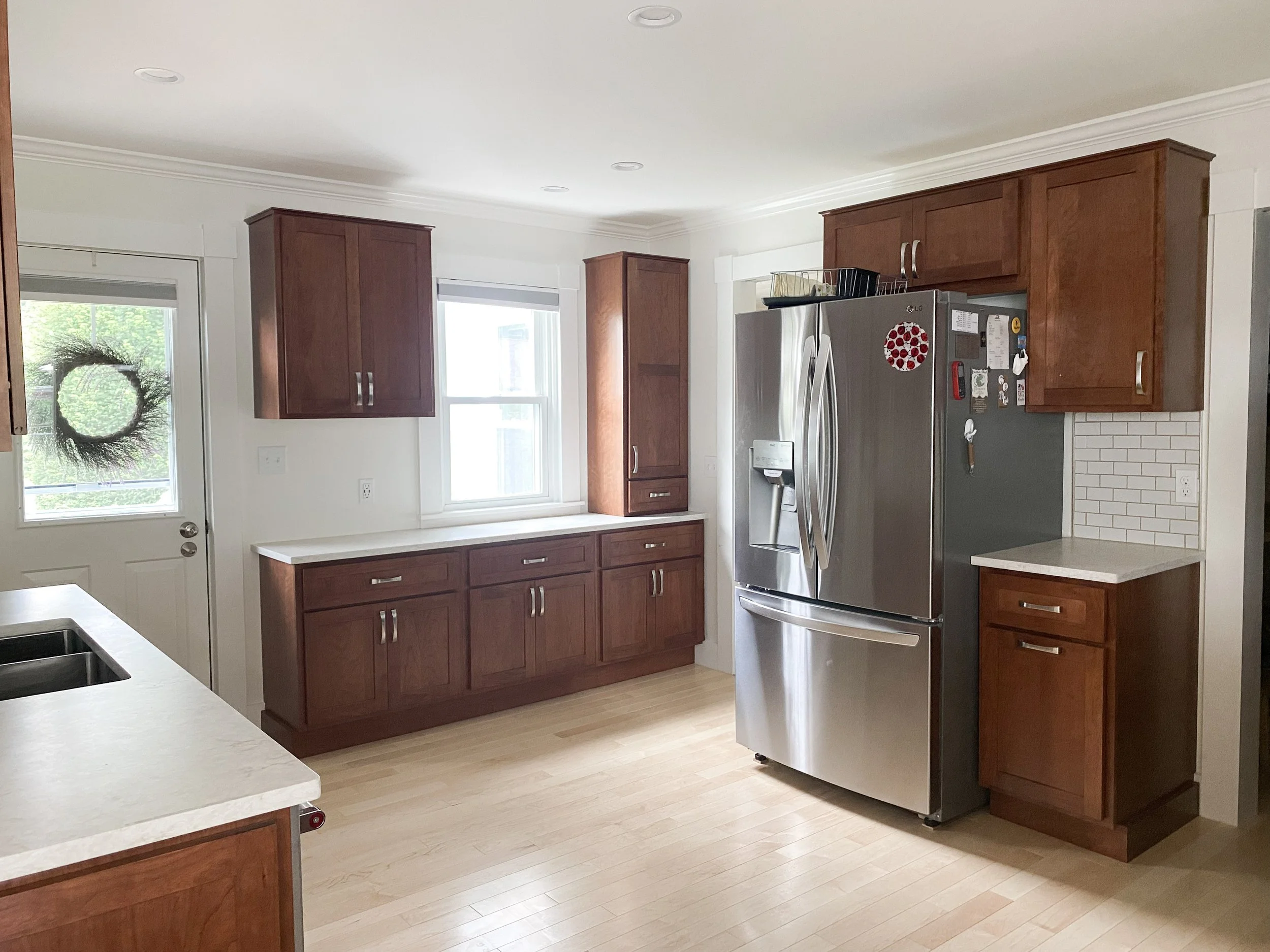 Kitchen with wooden cabinets, a stainless steel refrigerator, a white countertop, a window, and a door with a decorative wreath.