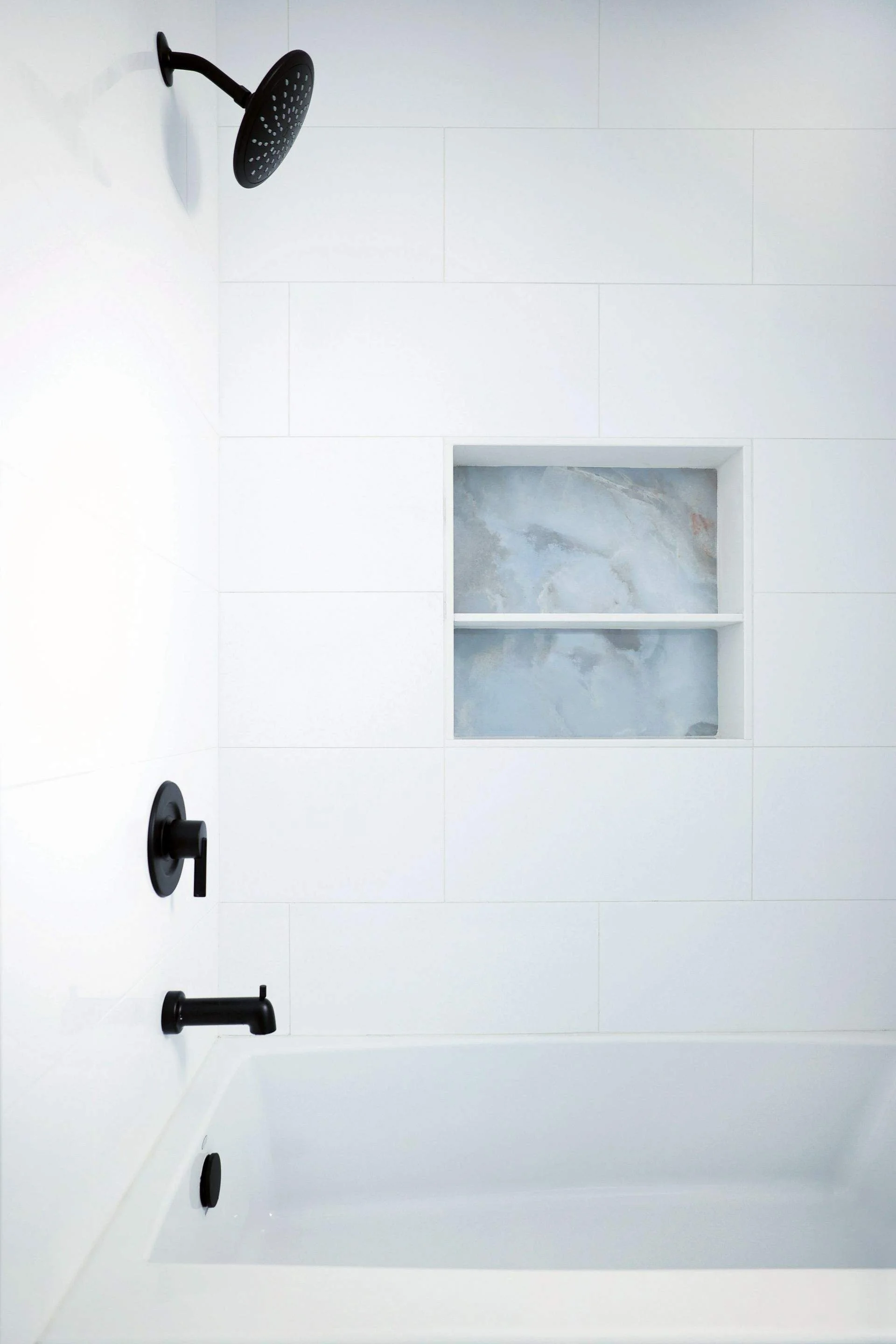 Top view of a modern white bathtub with black shower fixtures, including a round showerhead, a handle, and a spout, with a built-in niche in the tiled wall containing marble shelves.