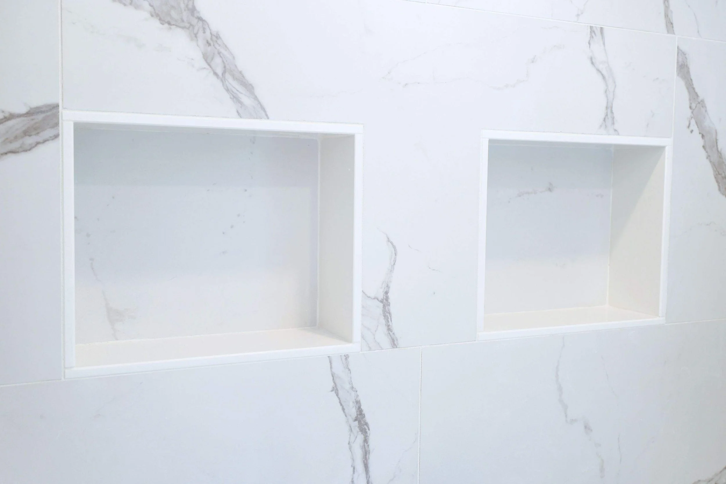 Two empty built-in rectangular shower niches in a marble tiled wall.