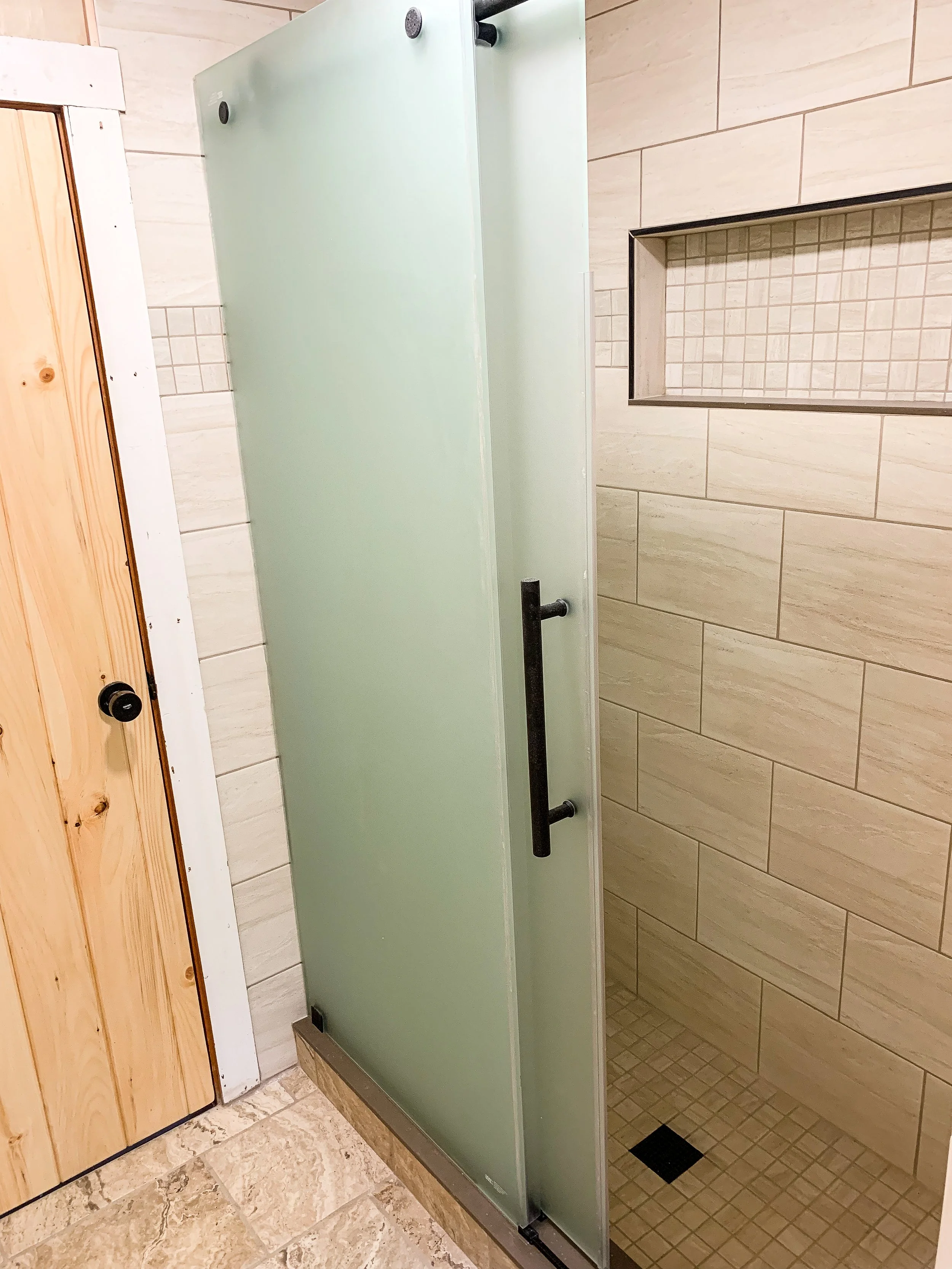 A frosted glass shower door with a black handle, partially opened, next to a tiled shower area in a bathroom.