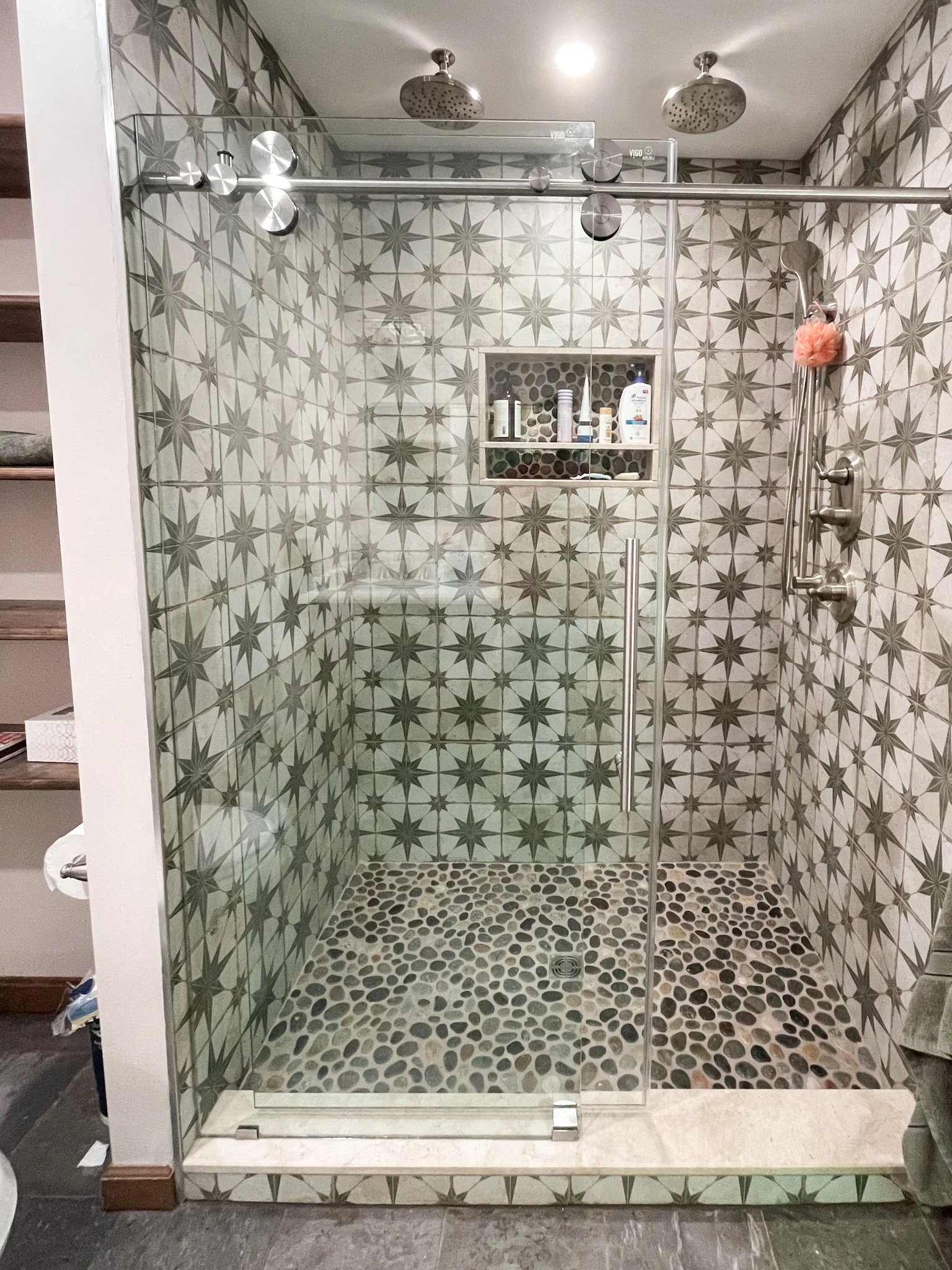 Walk-in shower with glass door, geometric star-patterned tile walls, pebble tile floor, two overhead showerheads, a built-in shelving niche with toiletries, a mounted hand-held showerhead with pink bath sponge, and a partial view of bathroom shelving and a towel.