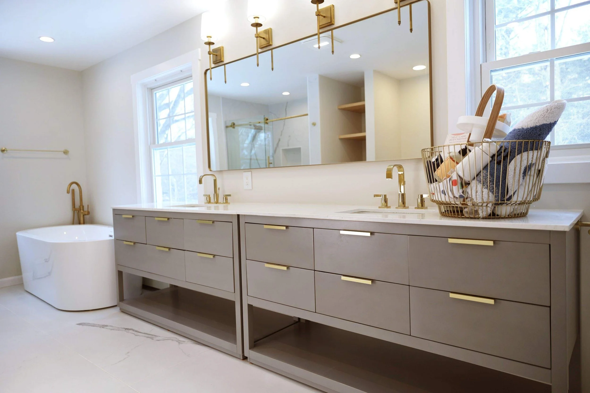 Modern bathroom featuring a double vanity with gray cabinets, gold fixtures, and a large mirror. There is a bathtub with gold fixtures on the left and a laundry basket with towels and supplies on the right. A window with a view of trees is above the countertop.