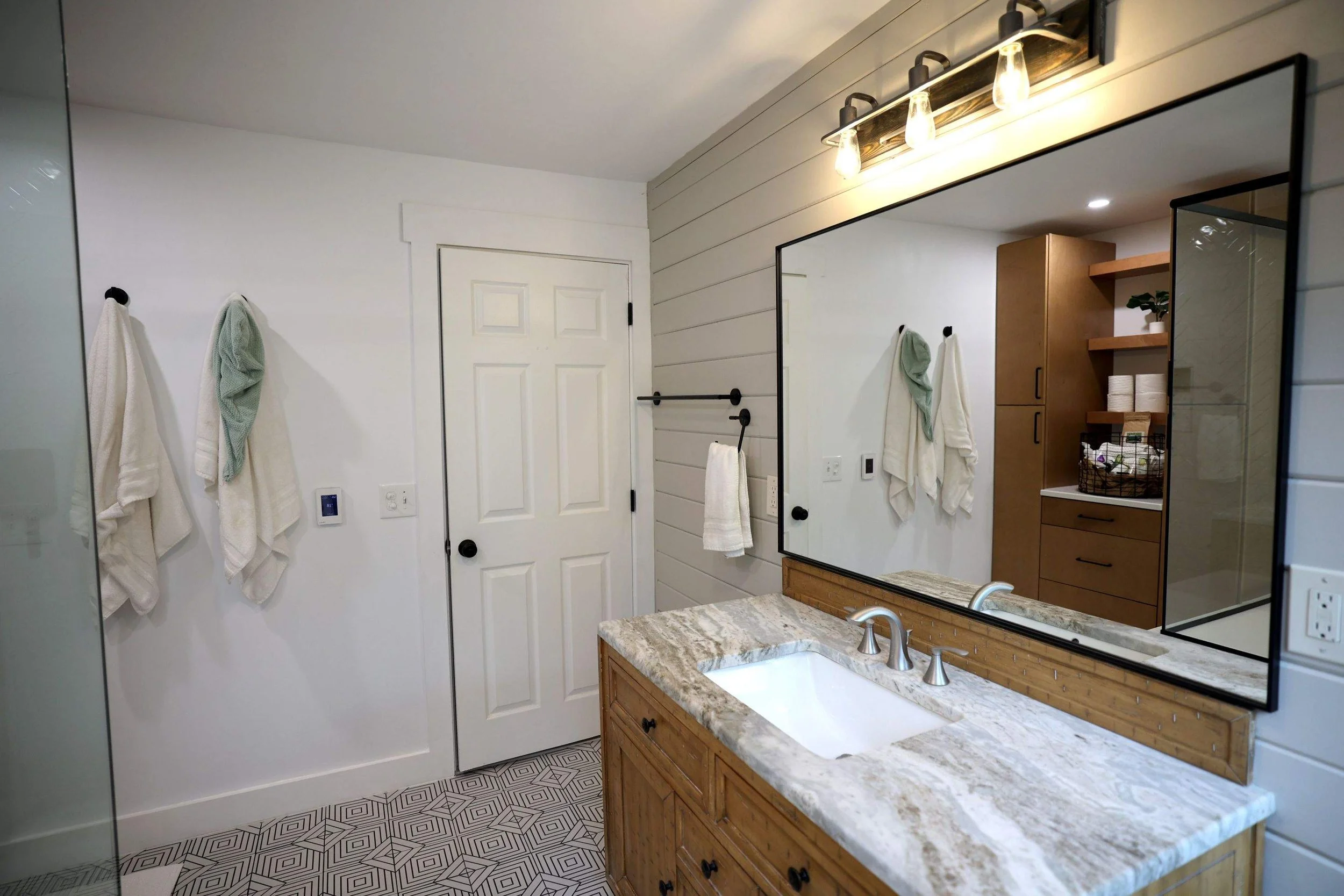 Bathroom with white walls, a wooden vanity with a granite countertop, a large mirror, hanging towels, and wooden shelves with rolled towels and decorative items.