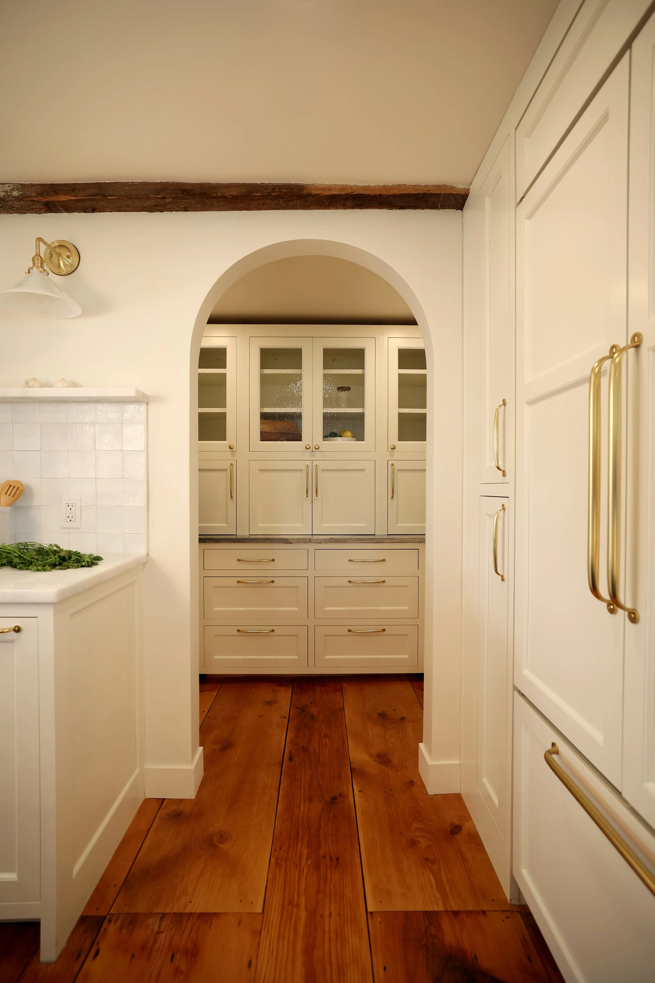 A view of a kitchen with white cabinetry, wooden floors, and an arched doorway leading to a pantry or storage area with glass-front cabinets.
