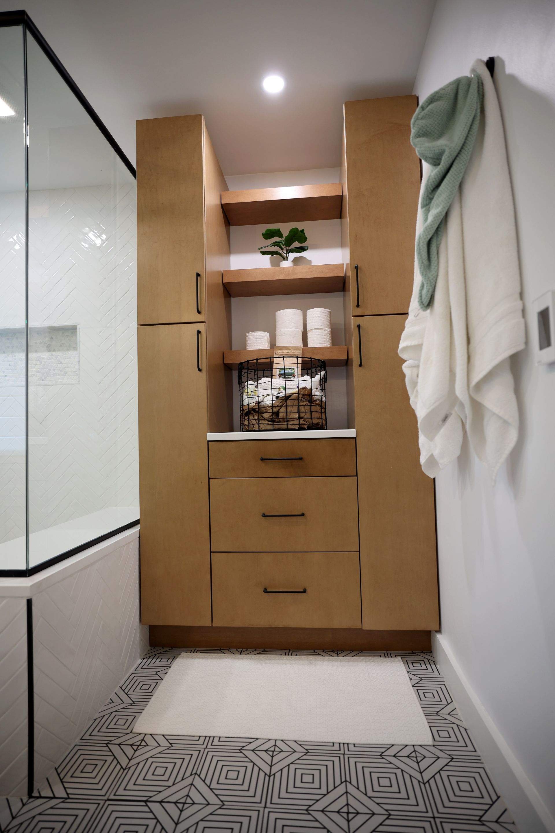 Wooden bathroom cabinet with shelves holding toilet paper rolls, a small plant, and a wire basket with towels, situated in front of a patterned floor and next to a glass shower enclosure.