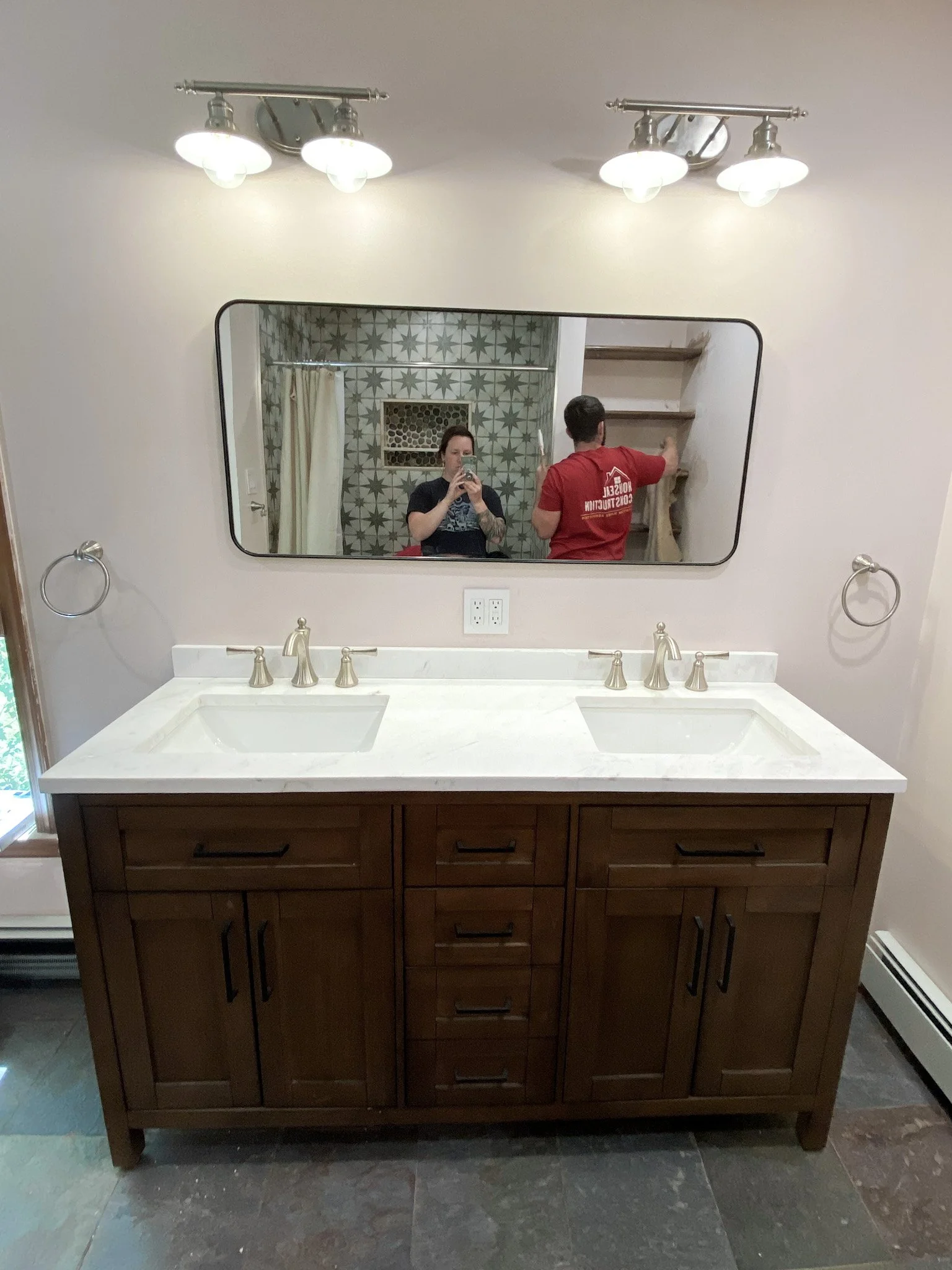 Bathroom vanity with two sinks, a large mirror, and overhead lights. Reflection shows two people, one taking a photo and the other working on a closet or cabinet.