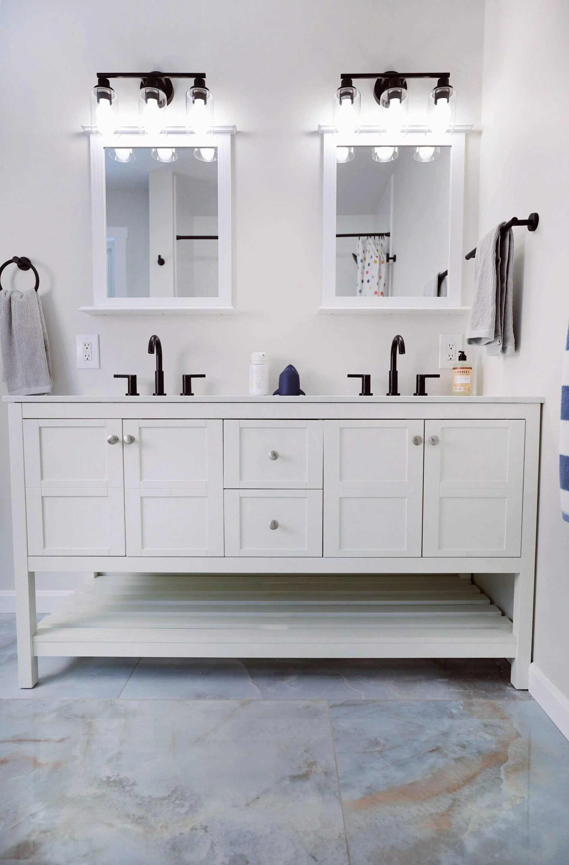 A white bathroom vanity with two sinks, two mirrors, and black fixtures. There are gray towels hanging on either side and a soap dispenser on the right side of the countertop.