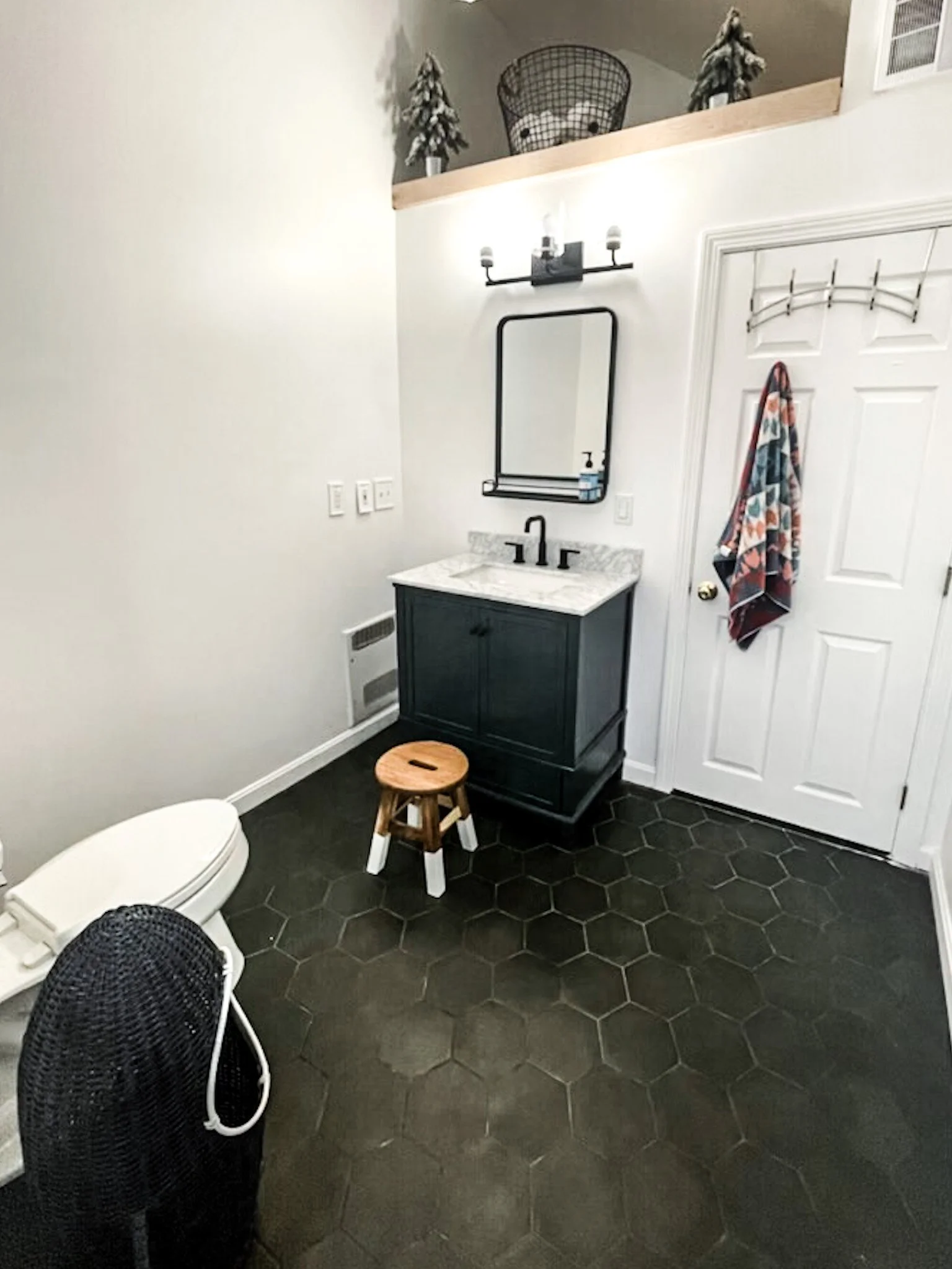 Small bathroom with black hexagon floor tiles, white walls, a white door with a colorful towel hanging, a black vanity with a marble top and a mirror above, a wooden stool, and a toilet. There are decorative plants and a wire basket on a ledge near the ceiling.