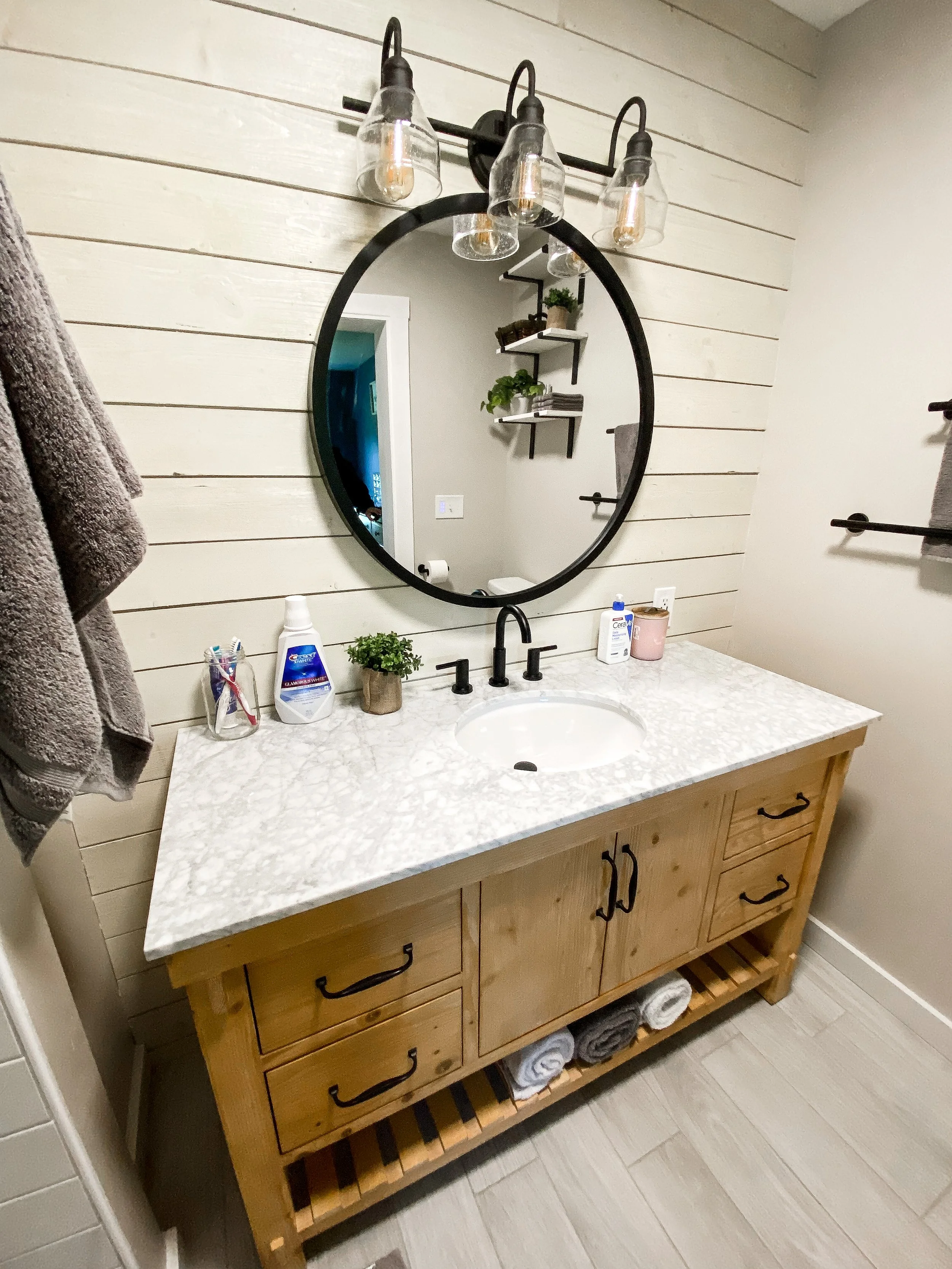 Bathroom vanity with a sink, black faucet, and oval mirror, against a white wood-paneled wall, with toiletries and towels on and below the wooden cabinet.