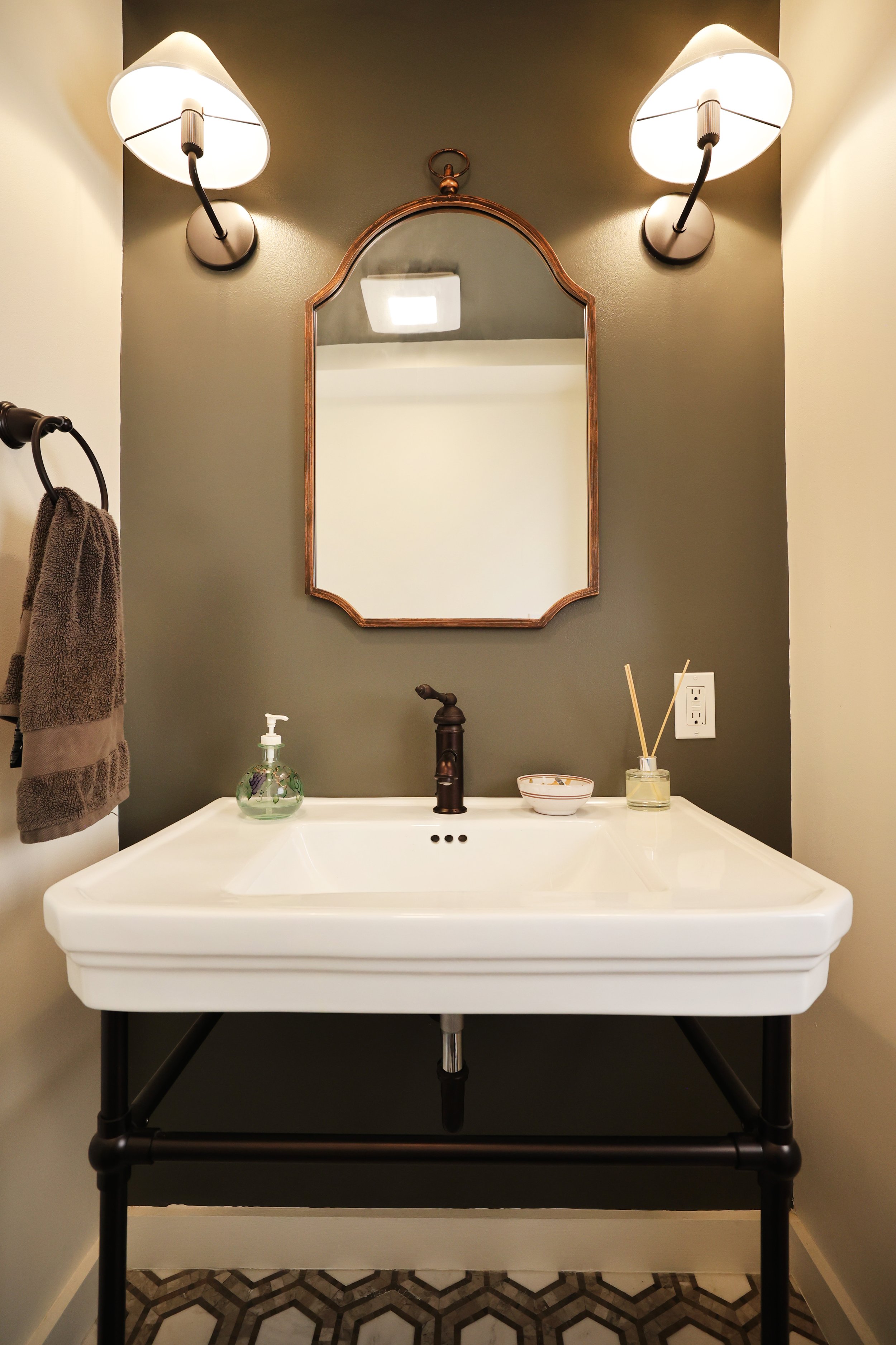 A bathroom vanity with a white sink, bronze faucet, and a mirror above, flanked by wall-mounted light fixtures. A hand soap dispenser, bowl, and reed diffuser are on the countertop, with a towel hanging on a towel ring to the left.